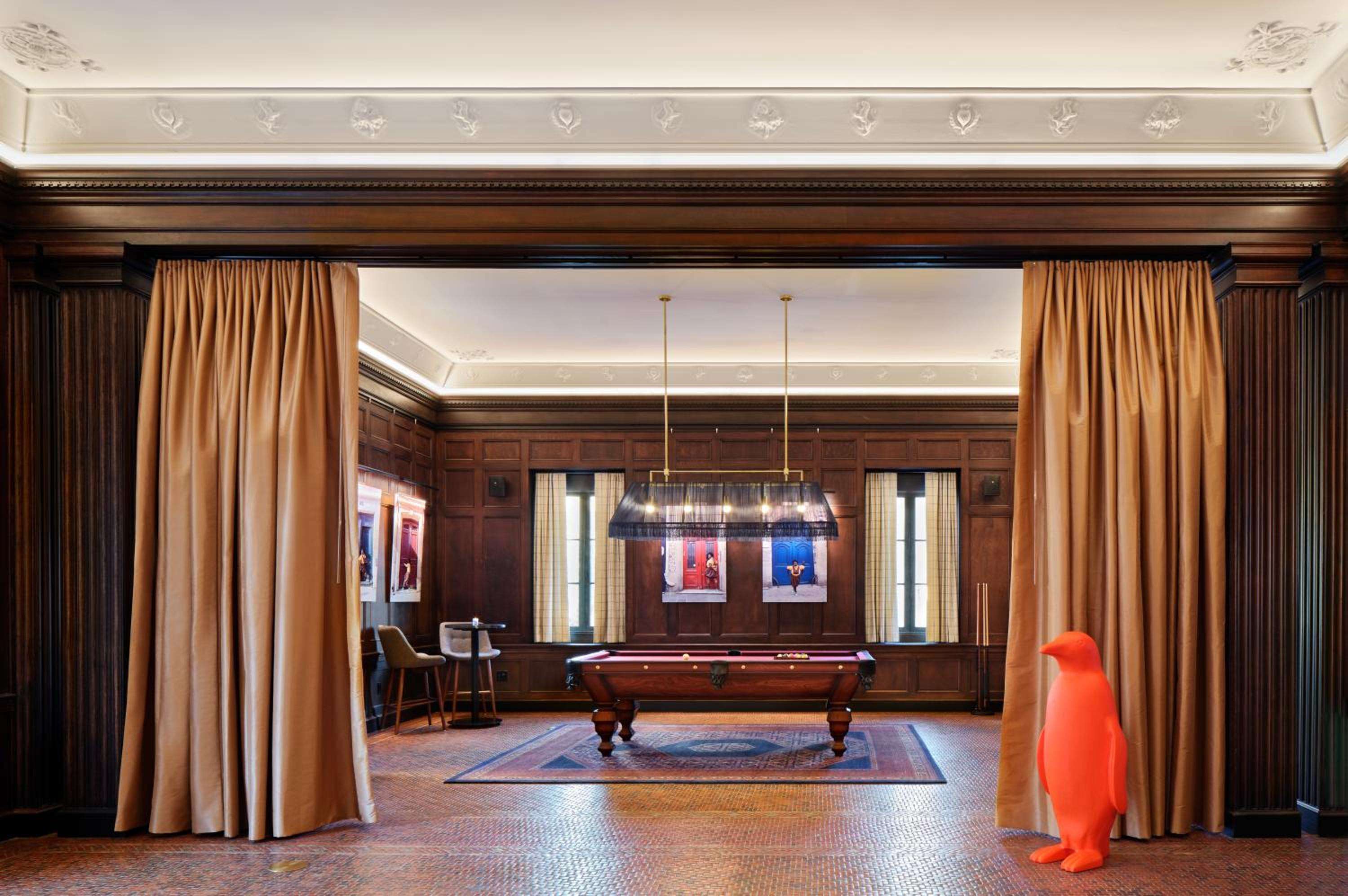 Rich mahogany paneling frames an elegant billiards room where contemporary art meets classic design, while a whimsical orange penguin sculpture adds unexpected artistic flair.