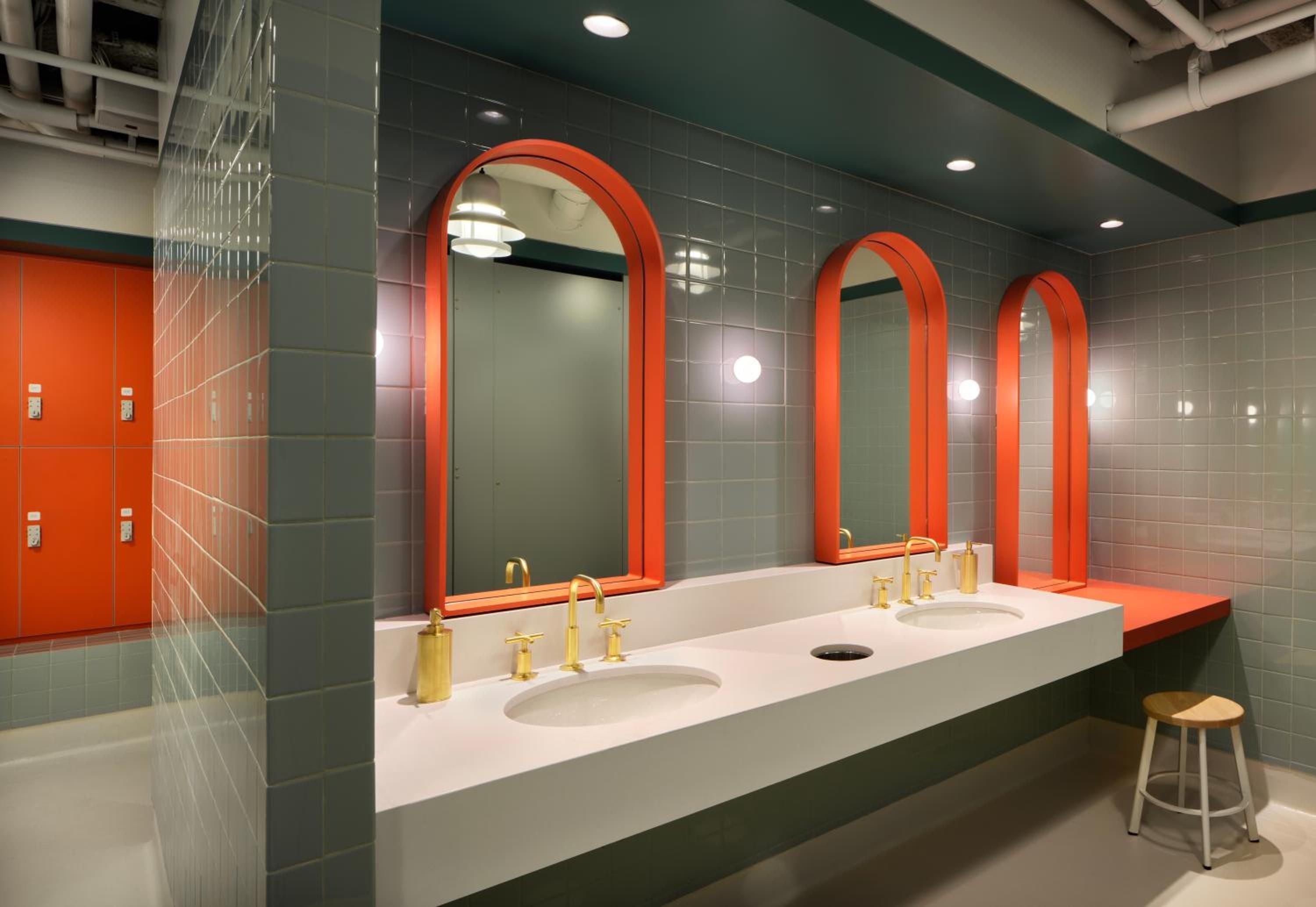 Bold orange-framed mirrors and brass fixtures create a playful yet sophisticated washroom experience, where contemporary art hotel design meets functional elegance in gleaming tile surfaces.