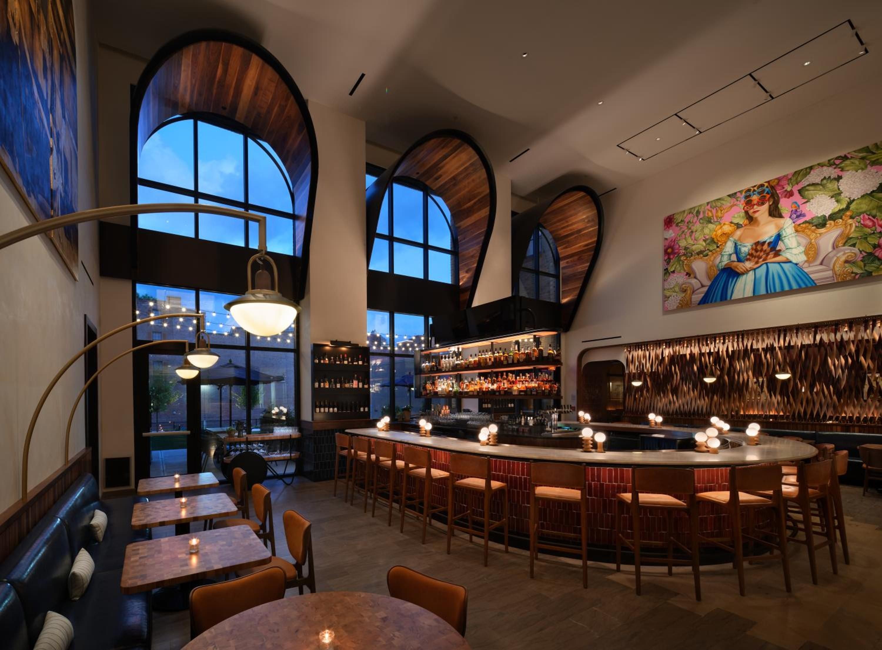 Curved banquette seating and an oval bar anchor this sophisticated dining space, where arched windows frame twilight views and warm pendant lights create intimate conversation pockets.