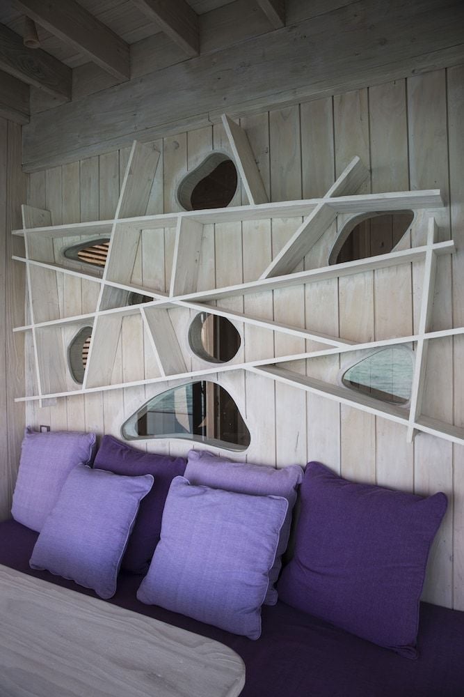 Weathered timber walls showcase whimsical porthole windows and sculptural white branches, while violet cushions invite afternoon contemplation in this imaginative overwater villa.