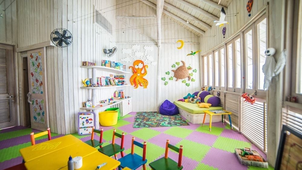 Colorful foam puzzle flooring and tiny rainbow chairs create a whimsical kids' club where little ones explore books and toys beneath soaring whitewashed timber beams.