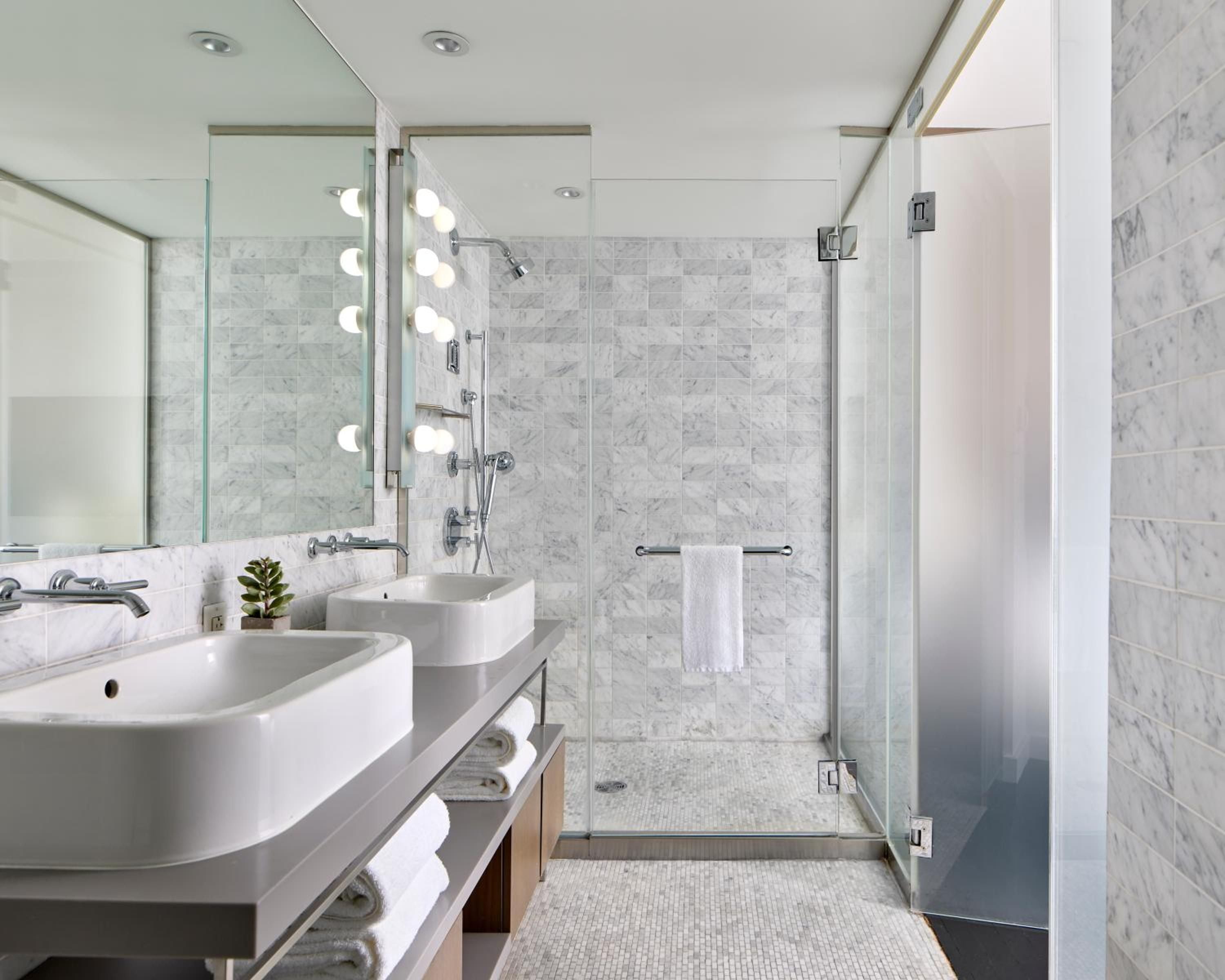 Carrara marble envelops the spa-like bathroom, where Hollywood-style vanity bulbs cast a warm glow over dual vessel sinks and frameless glass shower.