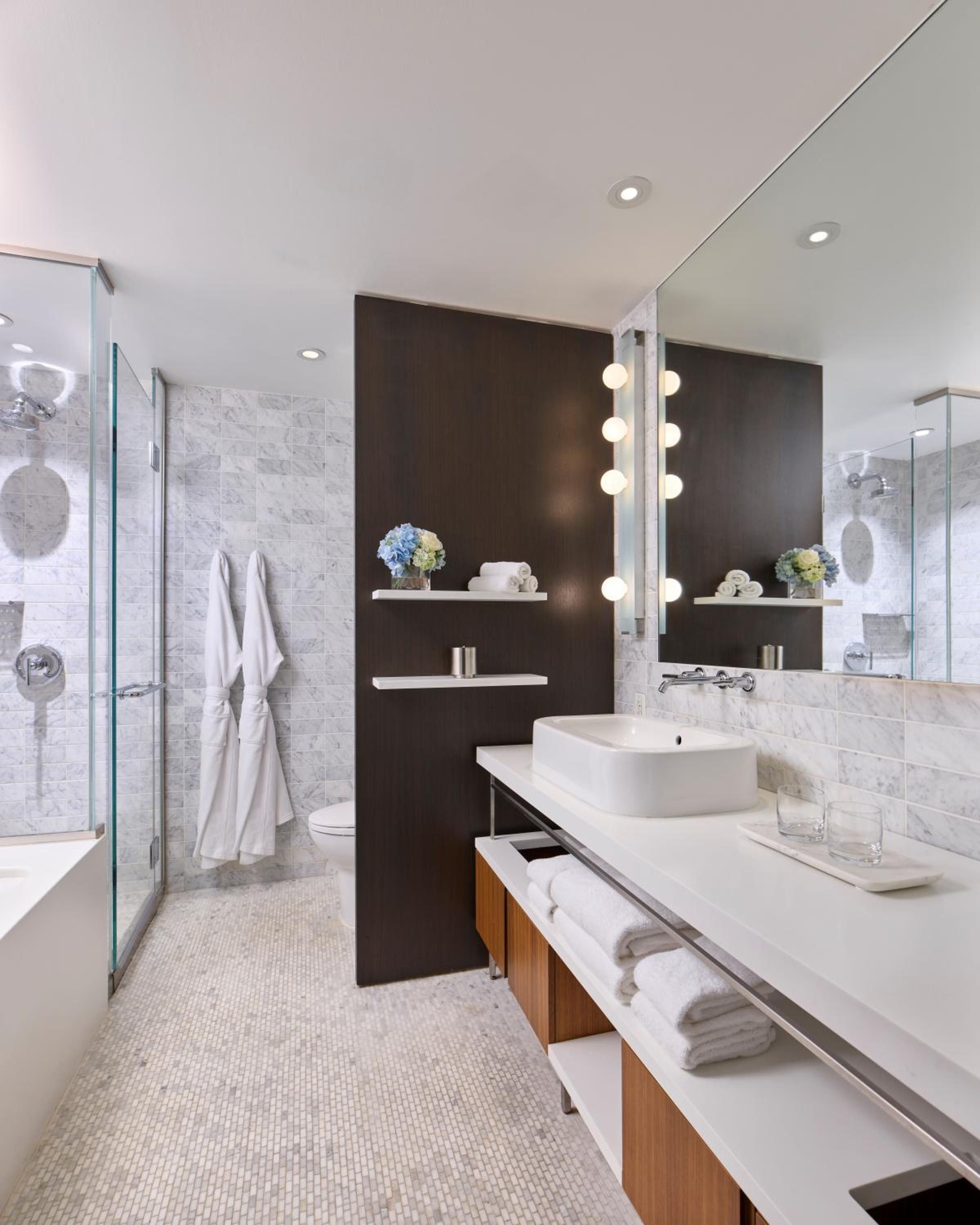 Warm wood vanities contrast against cool marble mosaics, while globe pendant lights create intimate illumination in this spa-like sanctuary with dual rainfall showers.