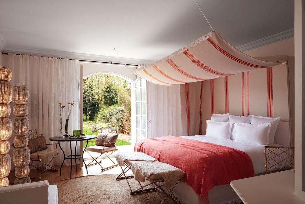 Coral-striped canopy drapes create intimate elegance above crisp white linens, while French doors frame verdant gardens beyond this romantic château suite.