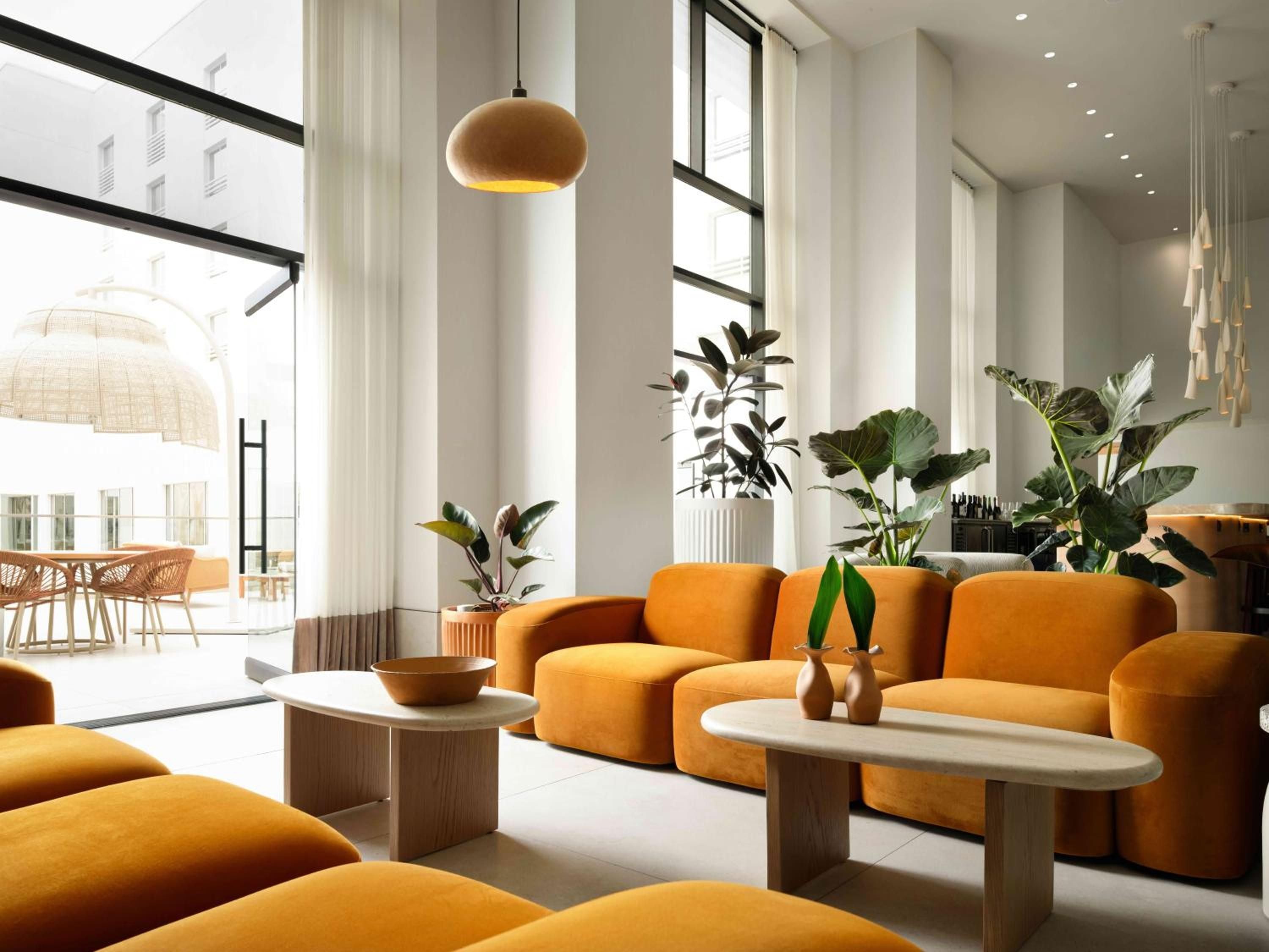 Warm amber pendant lights cast a golden glow over plush orange velvet seating, where tropical plants and sleek wood tables create an inviting modern sanctuary.