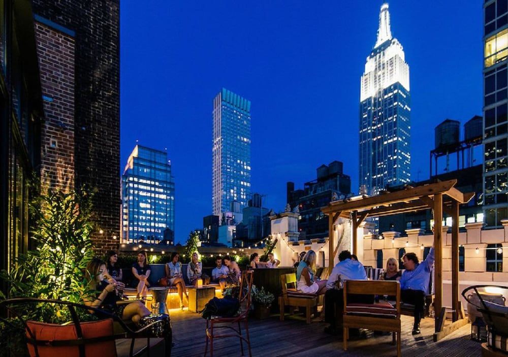 String lights twinkle above weathered wood seating as Manhattan's iconic skyline glows beyond this intimate rooftop retreat, where craft cocktails meet urban sophistication.