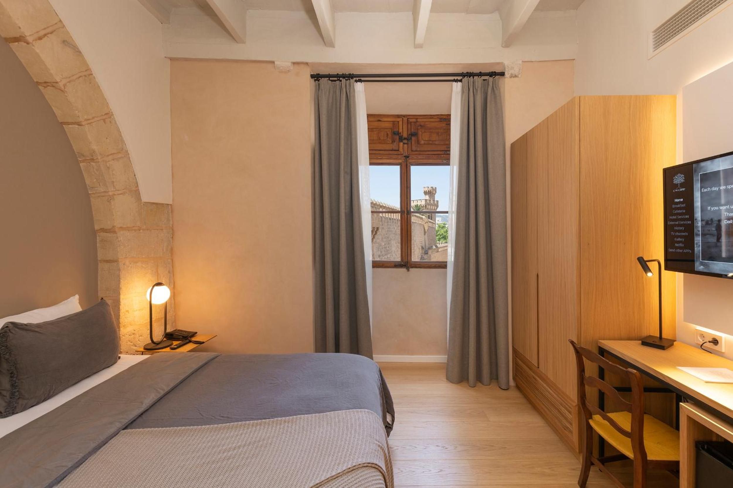 Stone archways and exposed beams frame this intimate suite, where warm wood furnishings complement historic Mallorcan architecture and glimpses of the cathedral beyond.