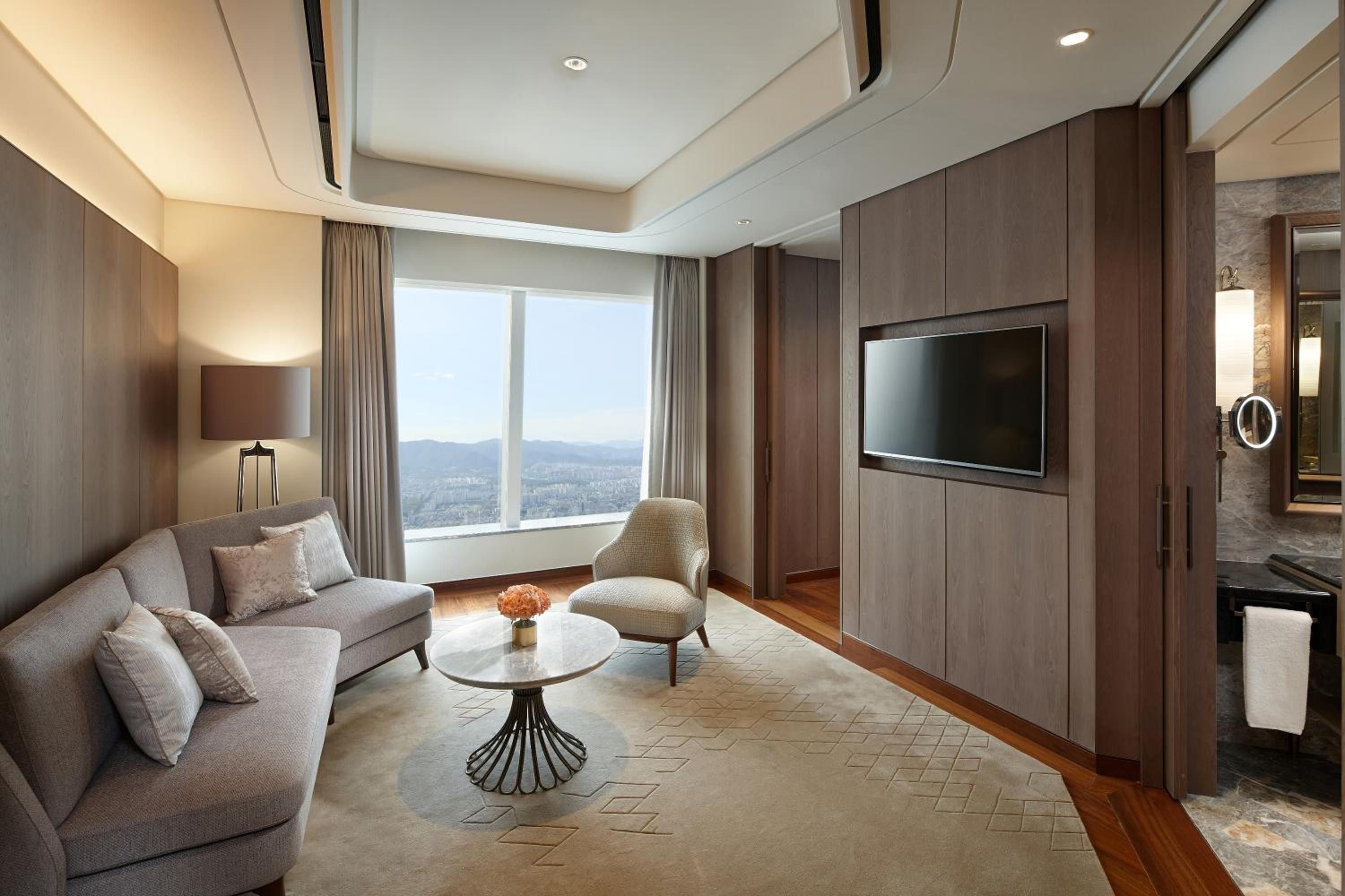 Floor-to-ceiling windows frame Seoul's sprawling cityscape from this sophisticated suite, where warm walnut paneling and plush gray furnishings create an elevated urban sanctuary.