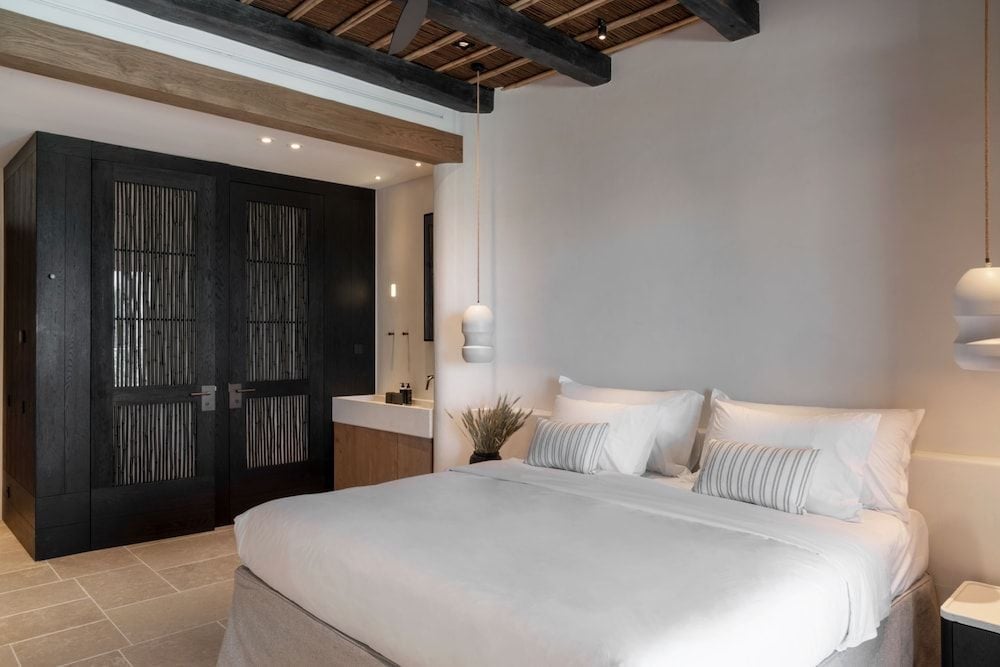 Exposed wooden beams frame this serene suite where charcoal-stained wardrobes and crisp white linens create a sophisticated harmony beneath Mykonos' traditional architecture.