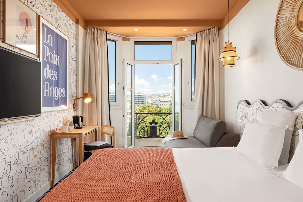 French doors frame Nice's Mediterranean cityscape from this thoughtfully appointed room, where coral throws warm minimalist furnishings and afternoon light dances across whitewashed walls.