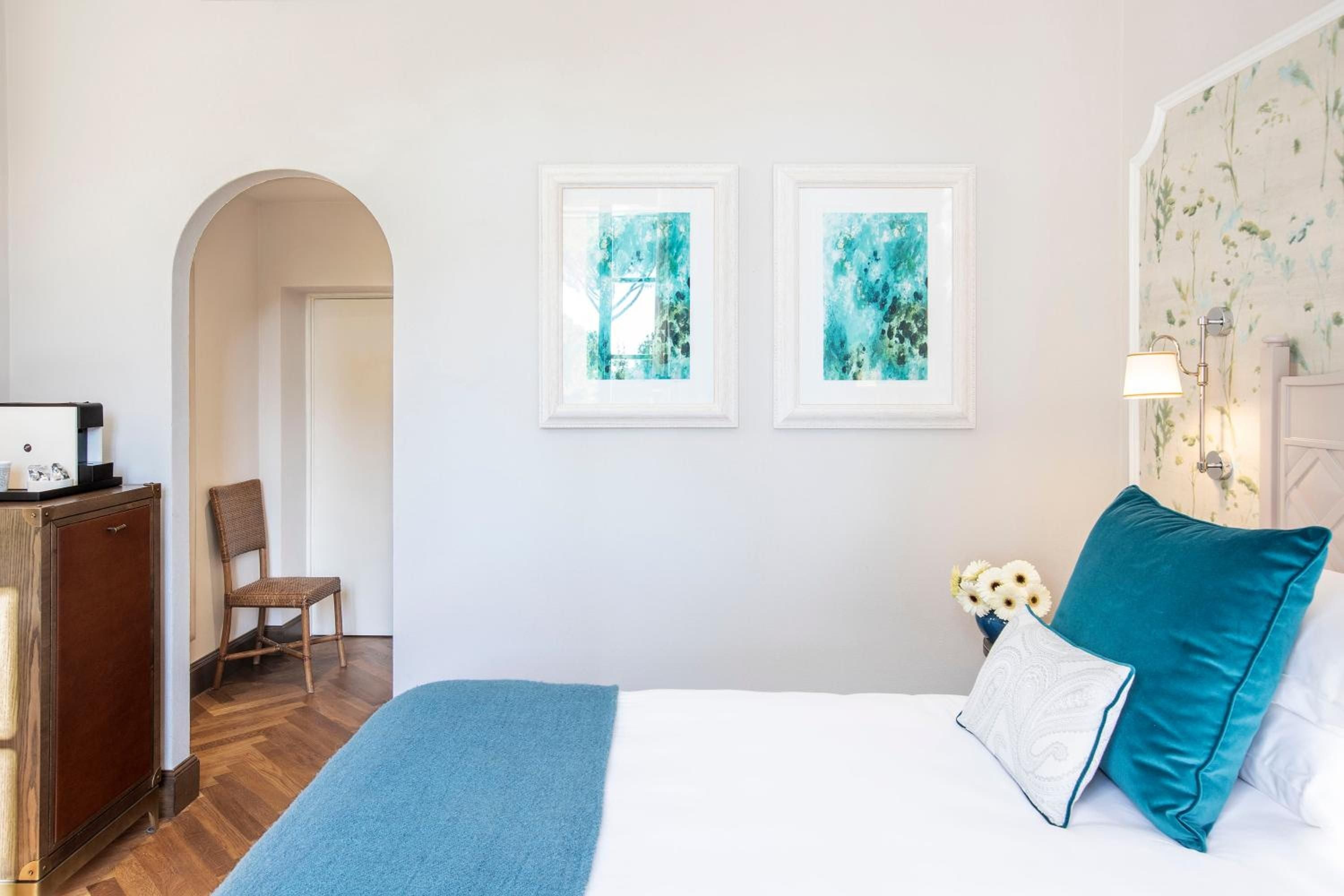 Soft morning light filters through botanical wallpaper while teal velvet pillows add Mediterranean flair to this thoughtfully appointed guest room with arched doorways and warm hardwood floors.