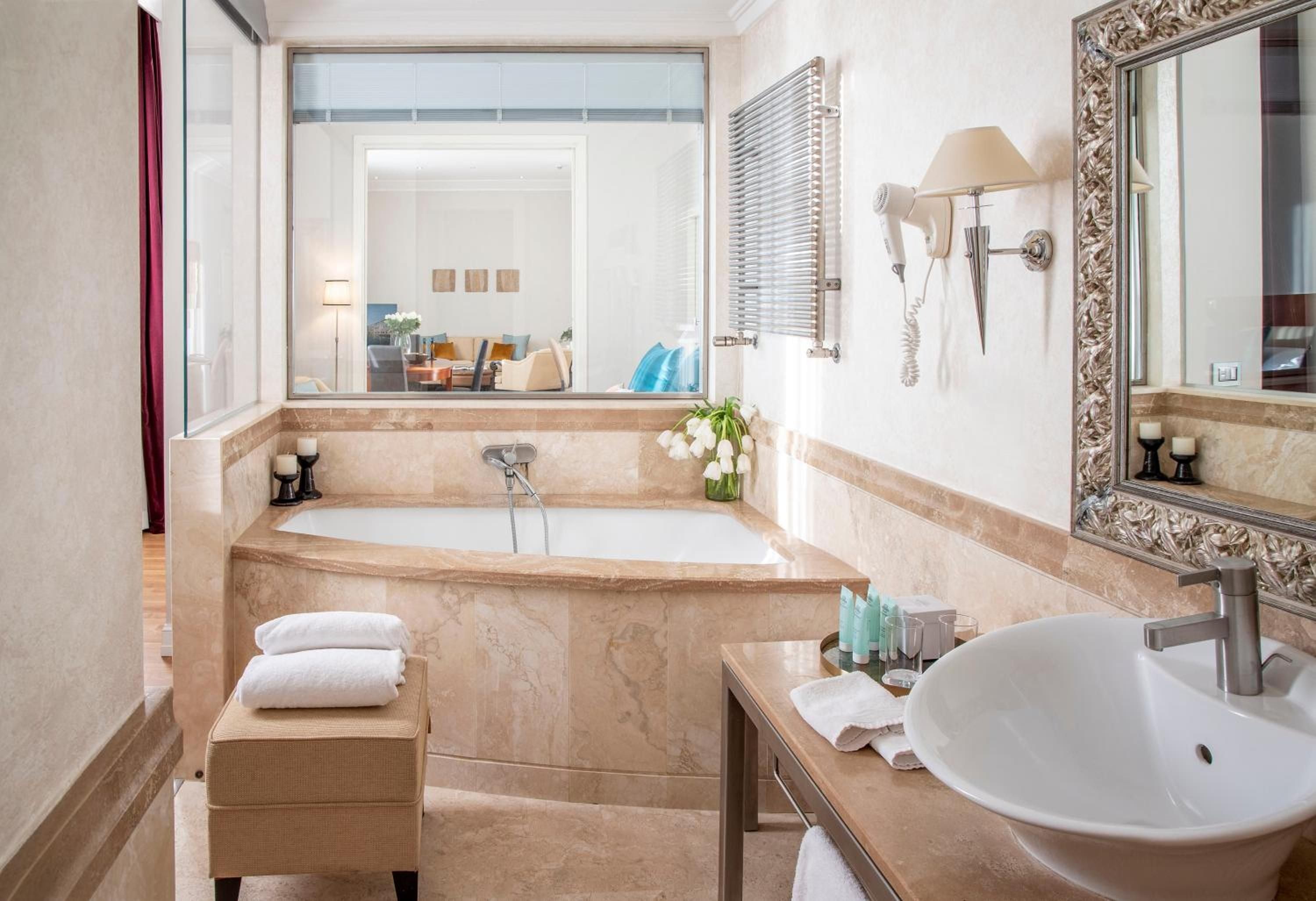 Creamy travertine surrounds the soaking tub where thermal waters await, while an ornate mirror reflects the sophisticated Italian craftsmanship defining every luxurious detail.