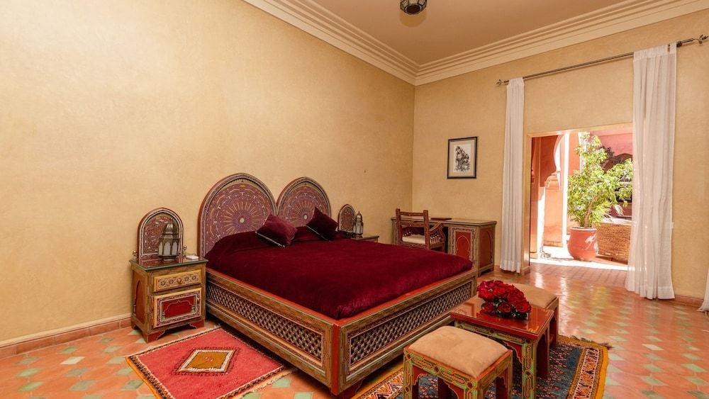 Intricate Moroccan woodwork frames the plush burgundy bedding while afternoon light streams across terracotta tiles, creating an intimate sanctuary of traditional craftsmanship.