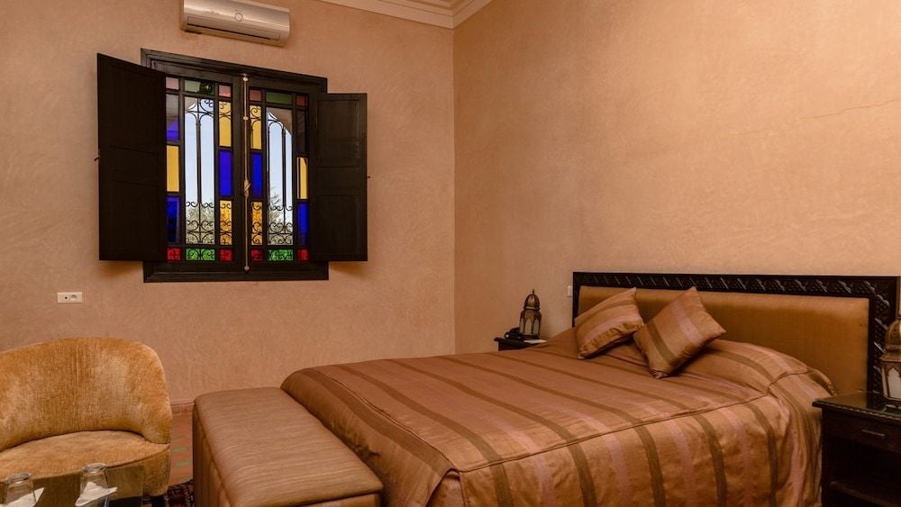 Warm terracotta walls embrace a guest room where vibrant stained glass shutters filter Moroccan light, casting jeweled patterns across striped linens and carved wooden furnishings.