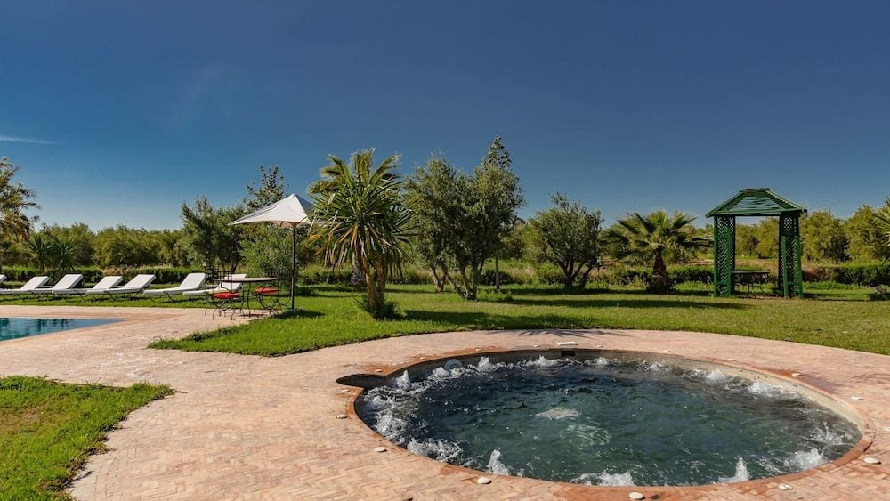 Stone-rimmed hot tub bubbles peacefully amid emerald lawns, while palm trees and a charming gazebo frame this tranquil Moroccan retreat under endless azure skies.