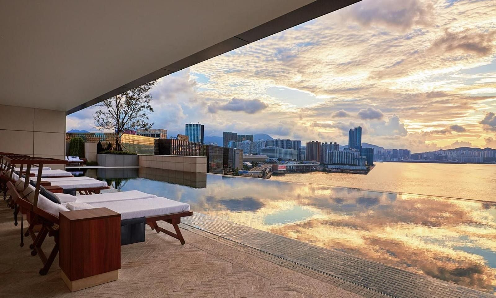 Golden hour transforms the infinity pool into liquid mirror, reflecting Victoria Harbour's glittering skyline while teak loungers await beneath the sculptural overhang.