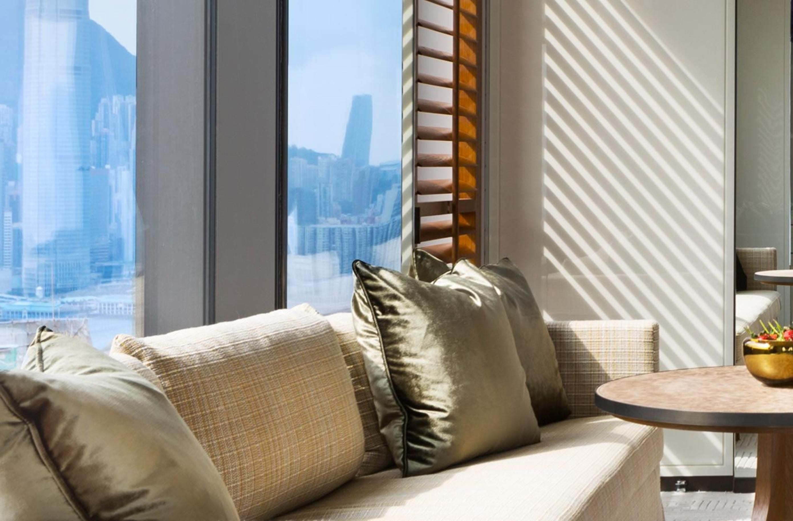 Afternoon light filters through custom blinds onto plush velvet cushions, while Hong Kong's glittering skyline unfolds beyond floor-to-ceiling windows in this serene suite retreat.