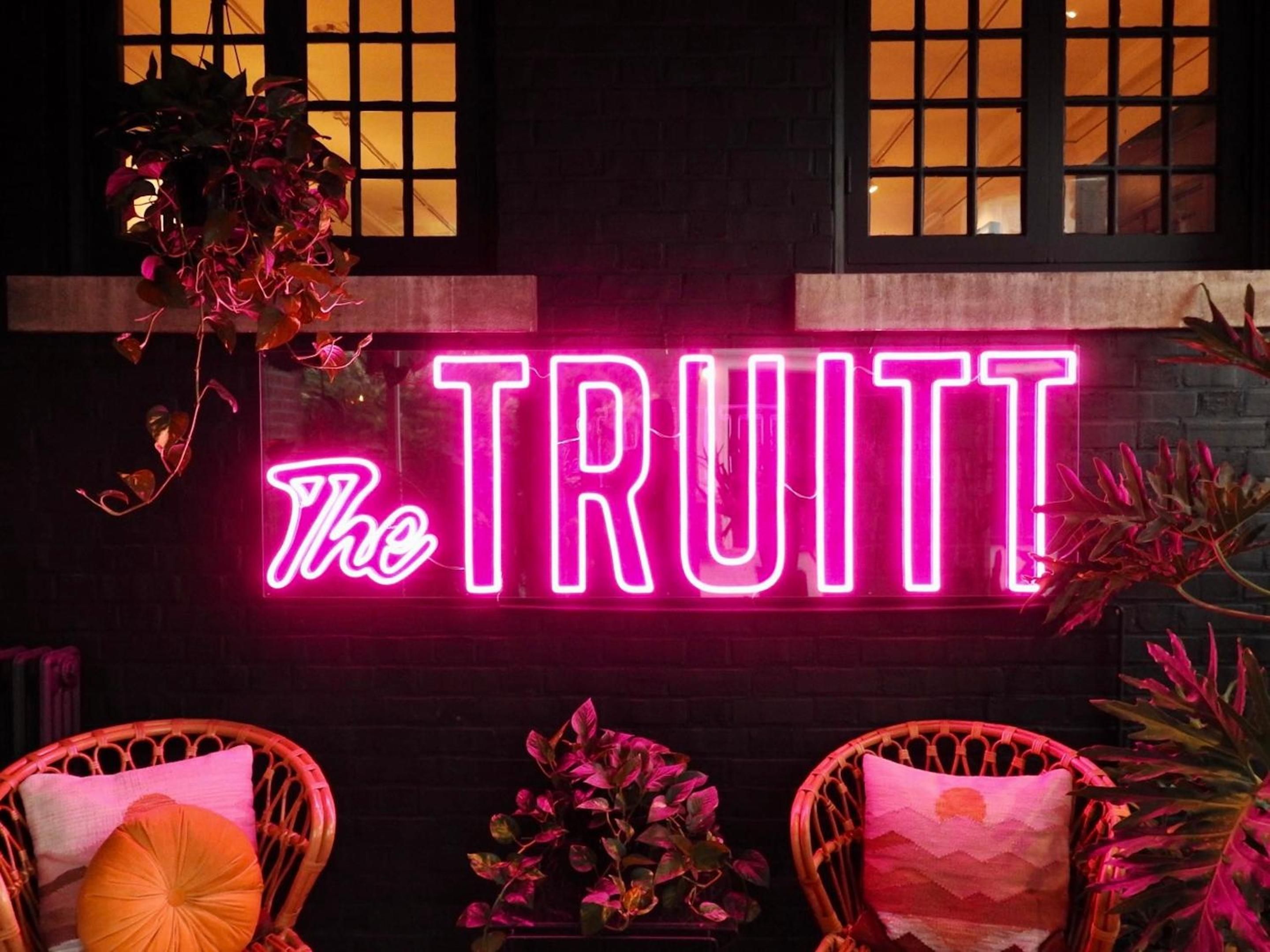 Pink neon light bathes wicker chairs and tropical plants in an intimate glow, while warm windows above create an inviting evening ambiance at this stylish drinking den.