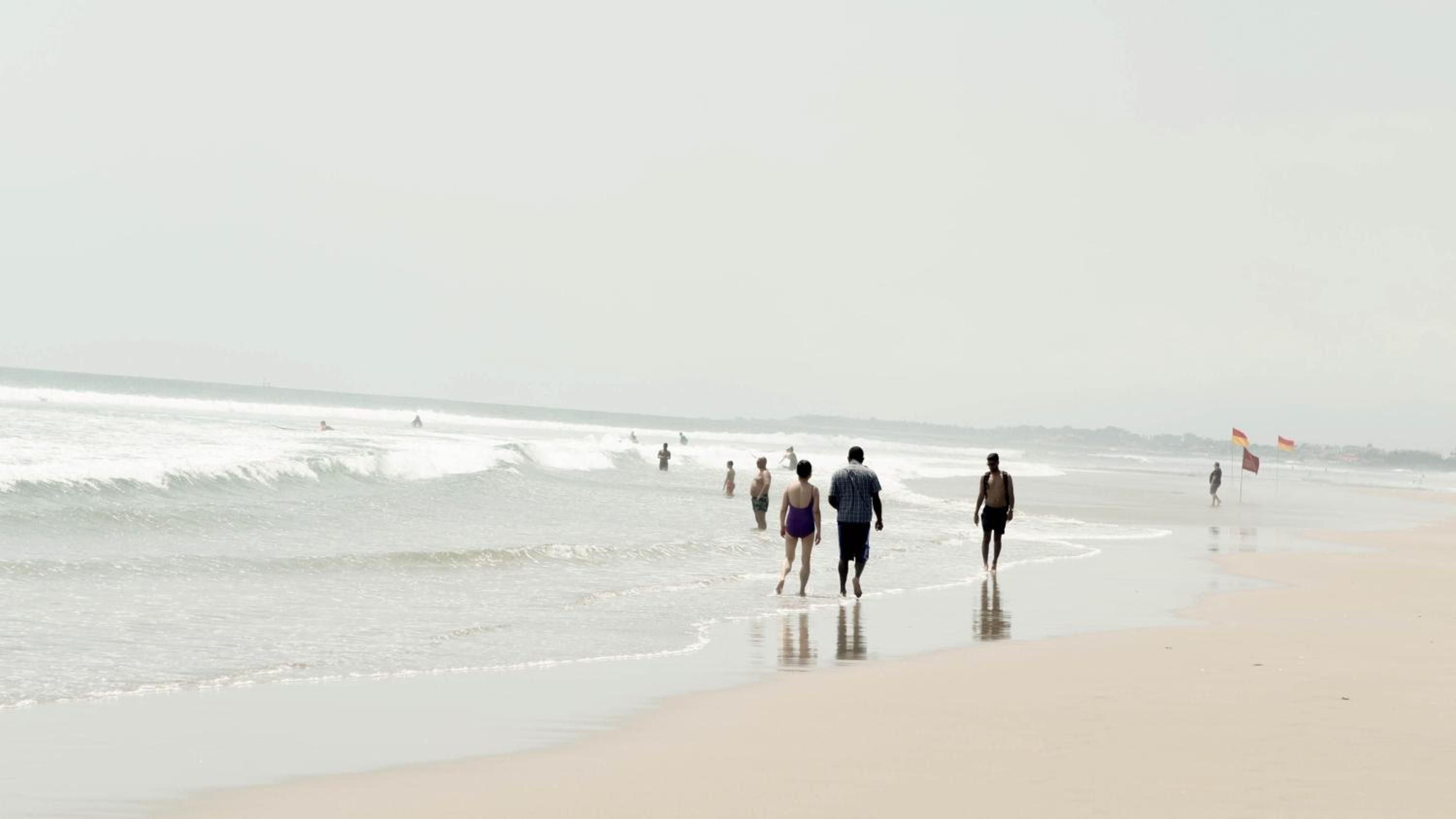 Misty morning waves roll onto pristine sand as beachgoers wade through gentle surf beneath an overcast sky, their silhouettes reflected in the wet shore.
