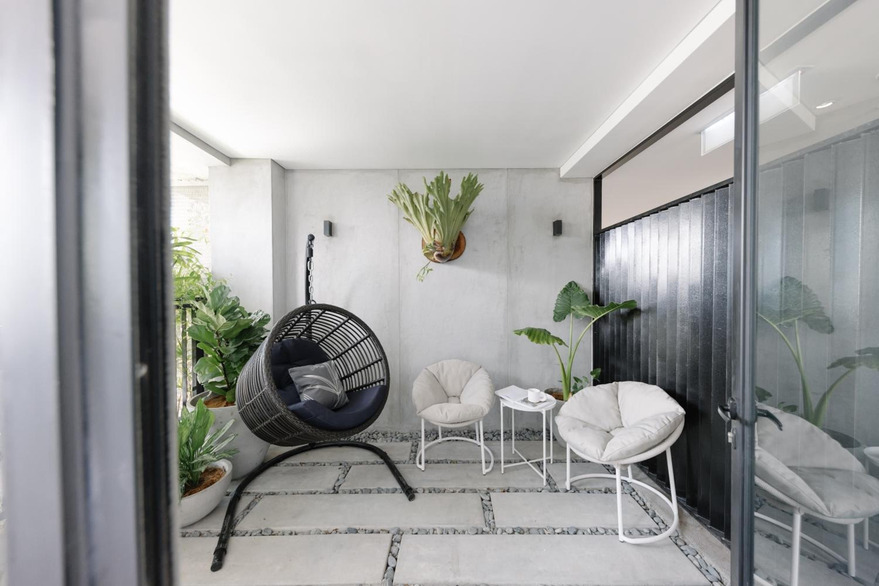 Sunlight filters through floor-to-ceiling glass onto a serene reading nook where suspended rattan chairs and tropical plants create an urban sanctuary of calm.