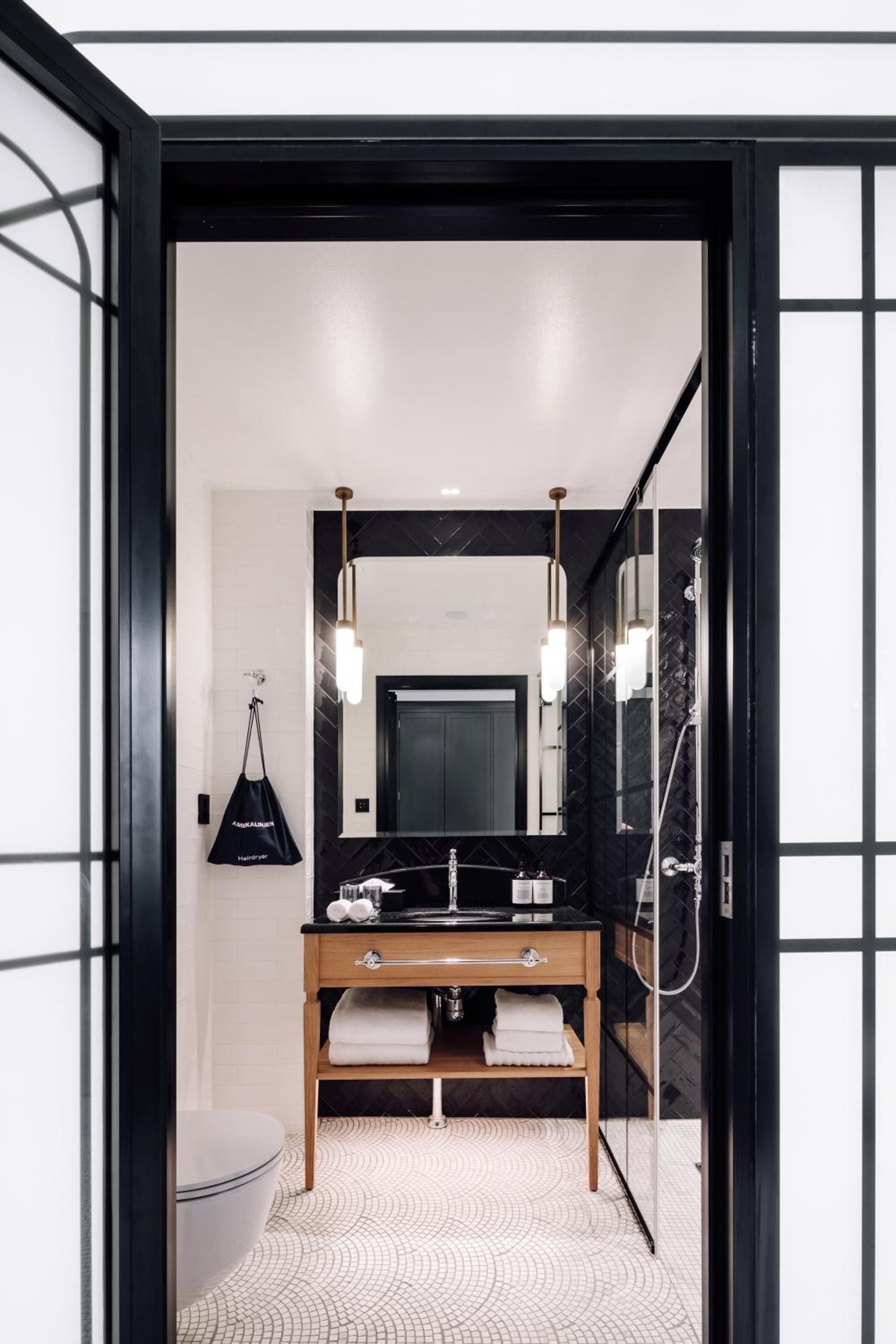 Black herringbone tiles and warm wood vanity create striking contrast in this thoughtfully designed bathroom, where brass pendant lights cast an intimate glow over marble surfaces.
