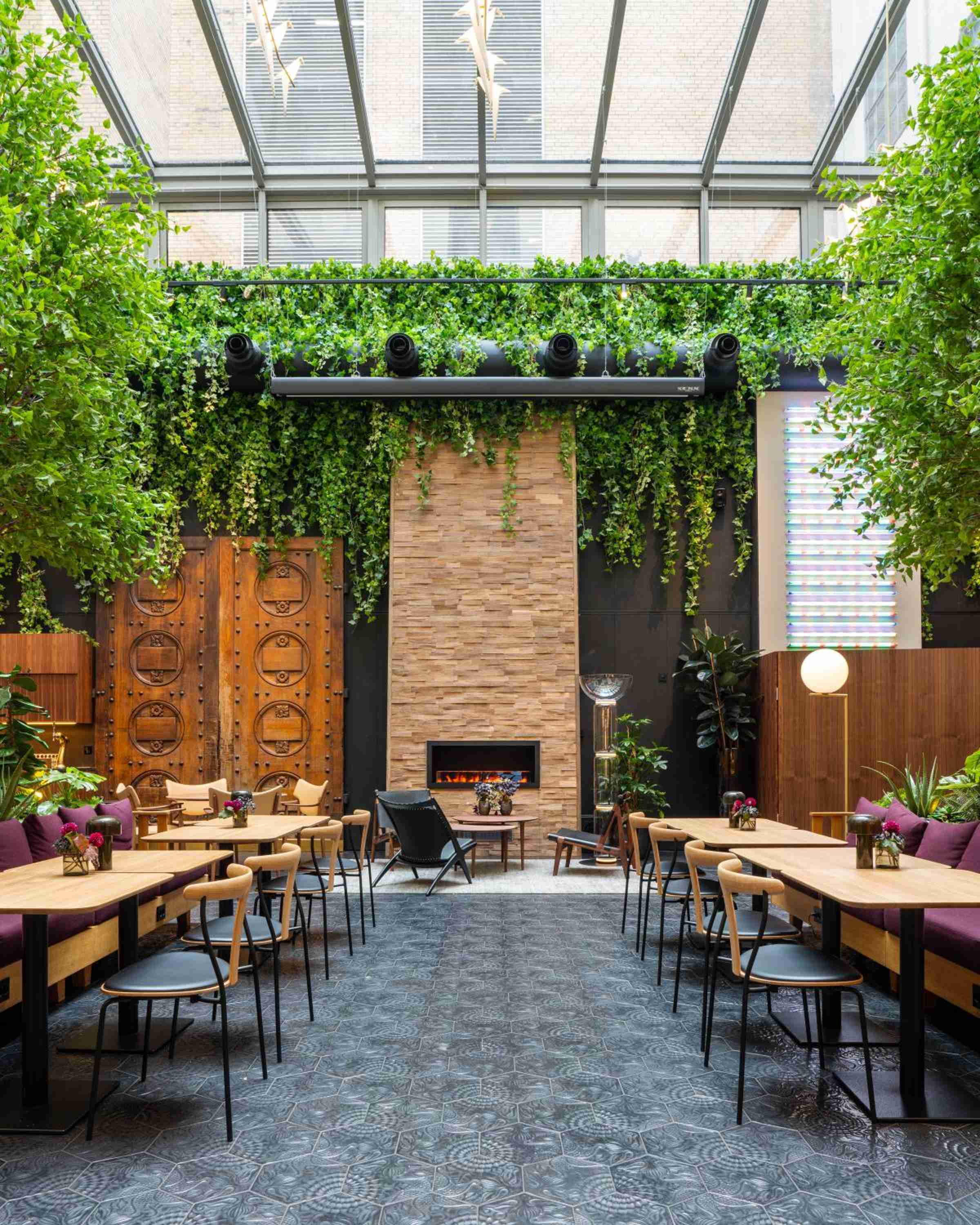 Cascading greenery frames the hotel's stunning restaurant courtyard, where natural light filters through glass ceilings onto warm wood furnishings and a glowing central fireplace.