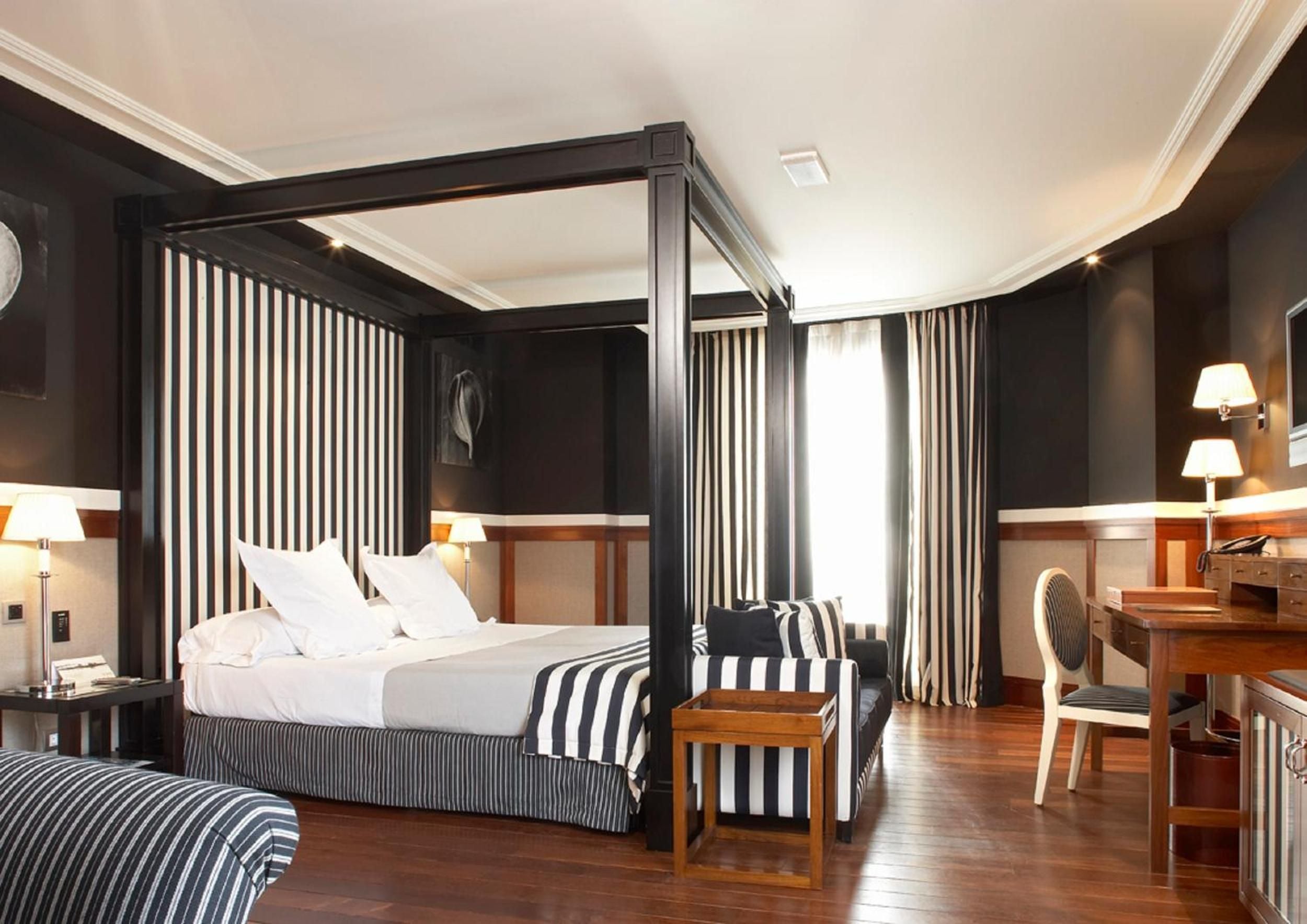 Rich mahogany floors gleam beneath a striking four-poster bed draped in bold black-and-white stripes, where maritime elegance meets contemporary Barcelona sophistication.