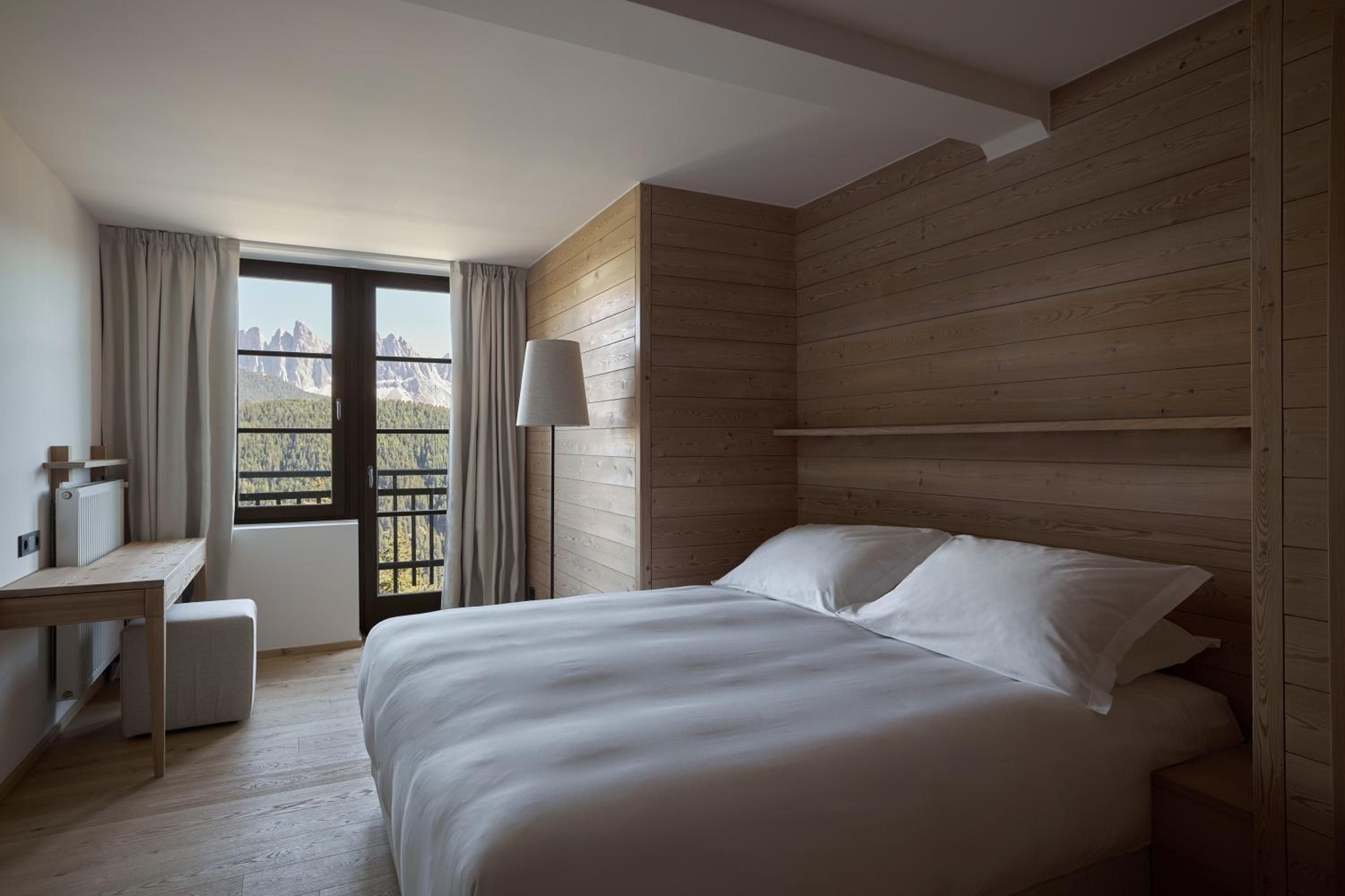 Natural larch wood envelops the minimalist suite, where crisp linens frame dramatic Dolomites peaks beyond floor-to-ceiling windows—alpine luxury distilled to its essence.