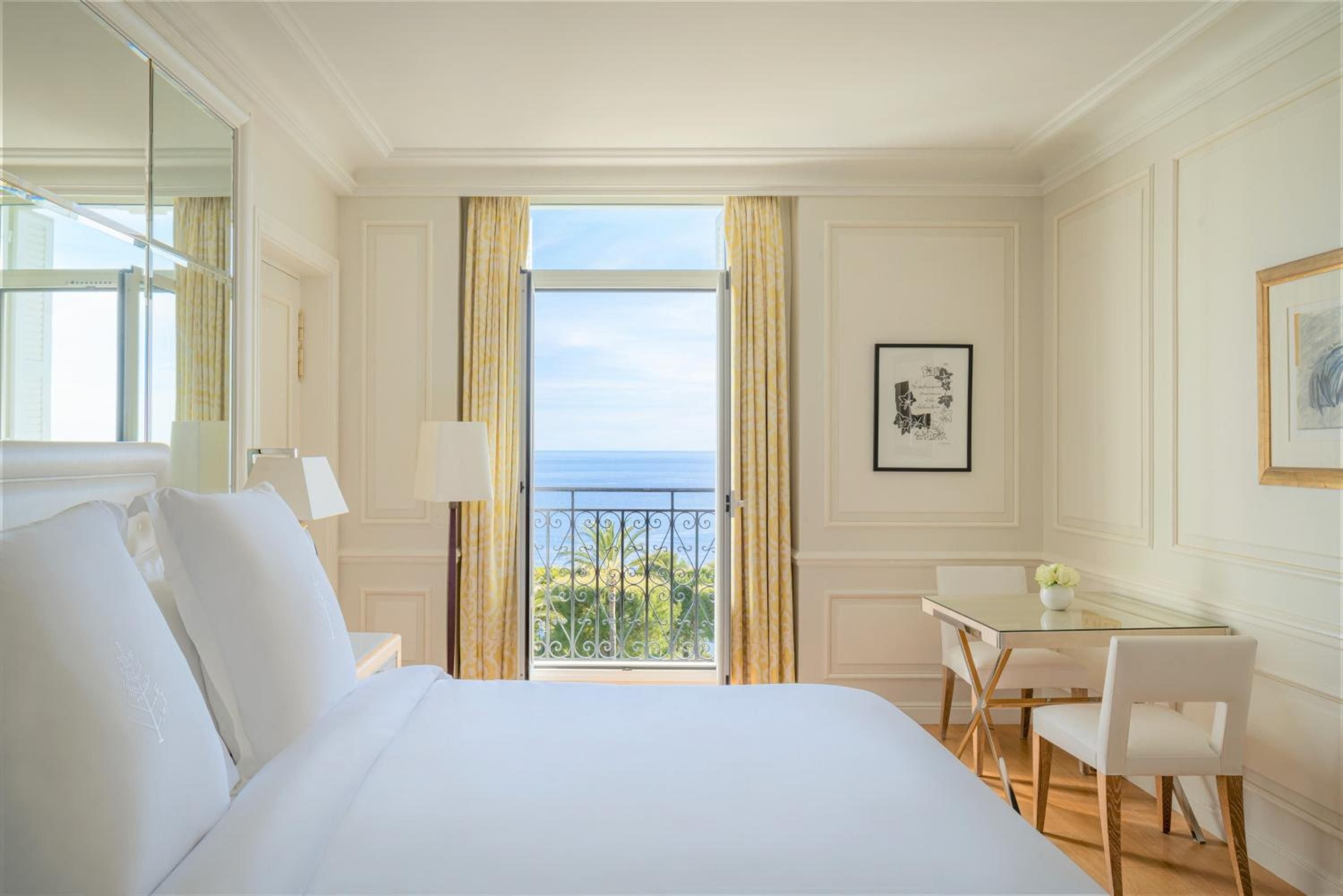 Crisp white linens frame panoramic Mediterranean vistas through French doors, where golden curtains filter Riviera light across polished parquet floors and classic Provençal elegance.
