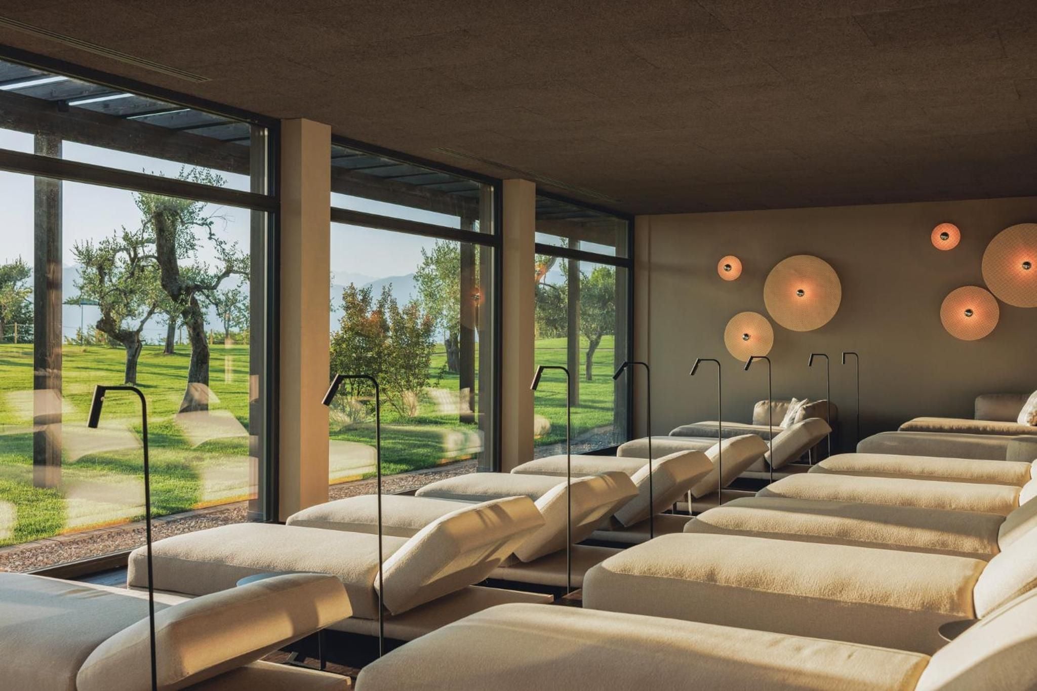 Heated relaxation loungers face olive groves through floor-to-ceiling windows, while sculptural ceiling lights cast a golden glow across this serene wellness sanctuary.