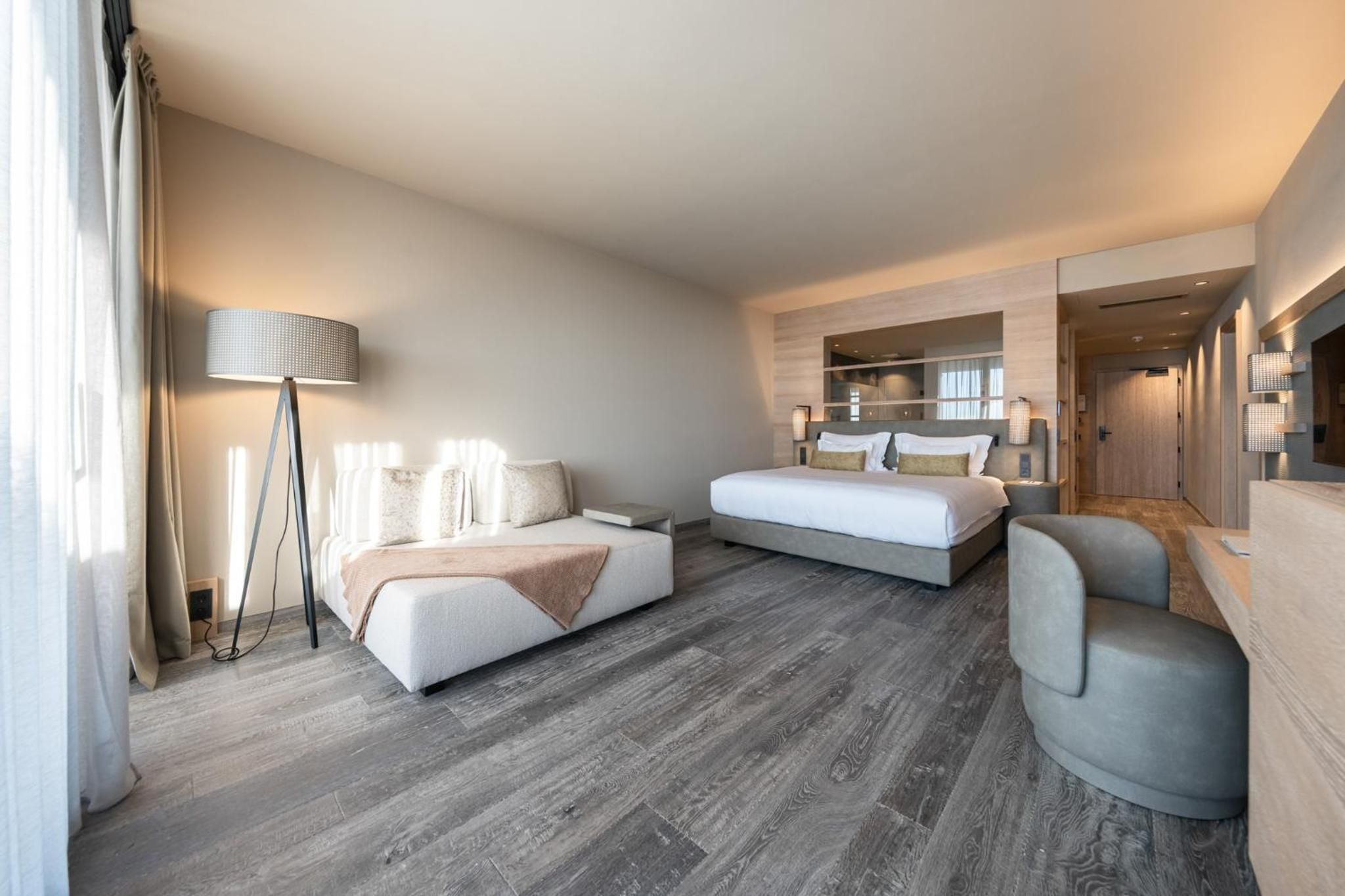 Weathered wood floors and neutral linens create a serene sanctuary where floor-to-ceiling windows bathe the spacious suite in golden light.