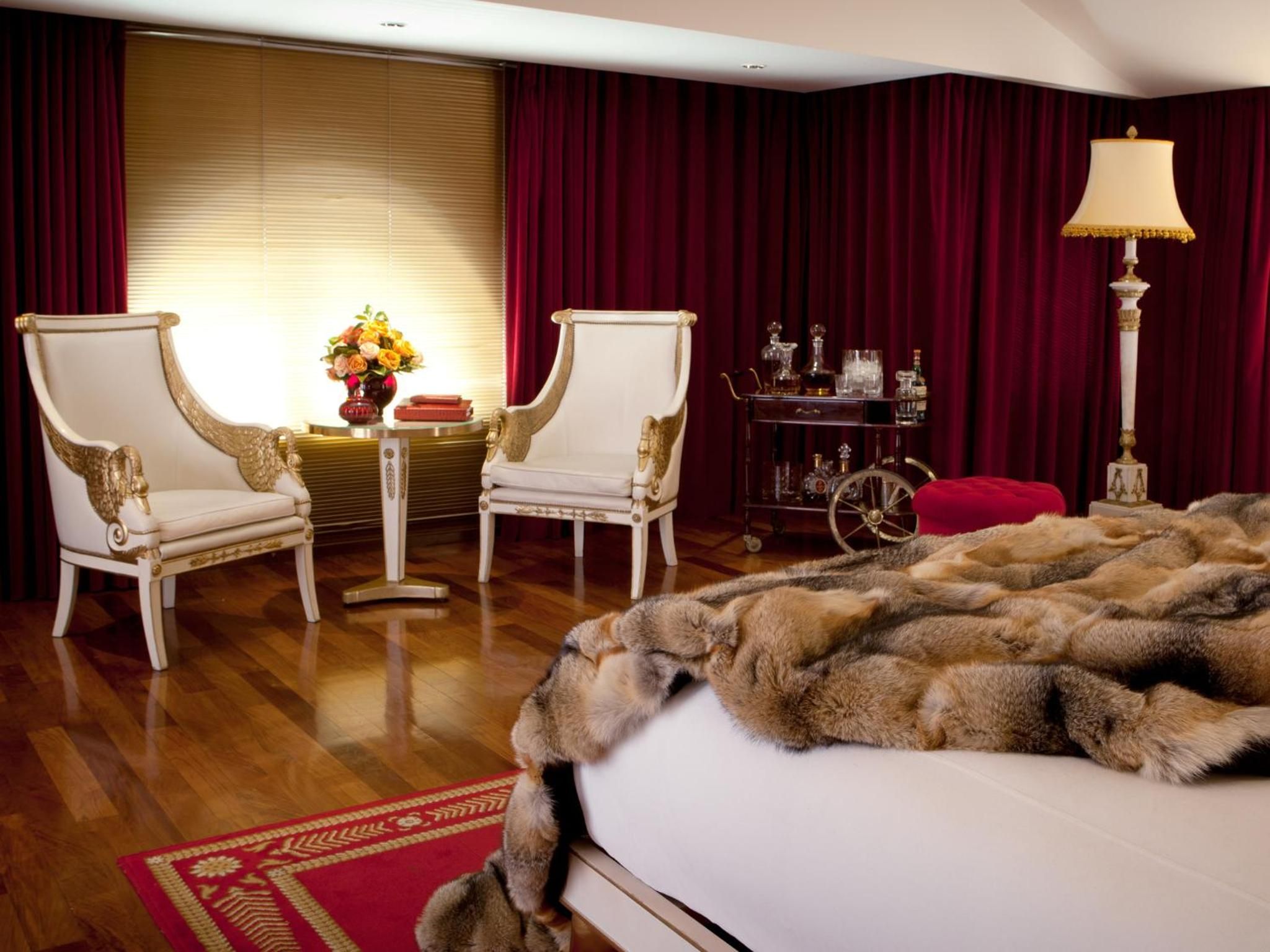 Sumptuous fur throws cascade across pristine linens while gilded Empire chairs create an intimate seating area beneath dramatic burgundy velvet curtains and golden lamplight.