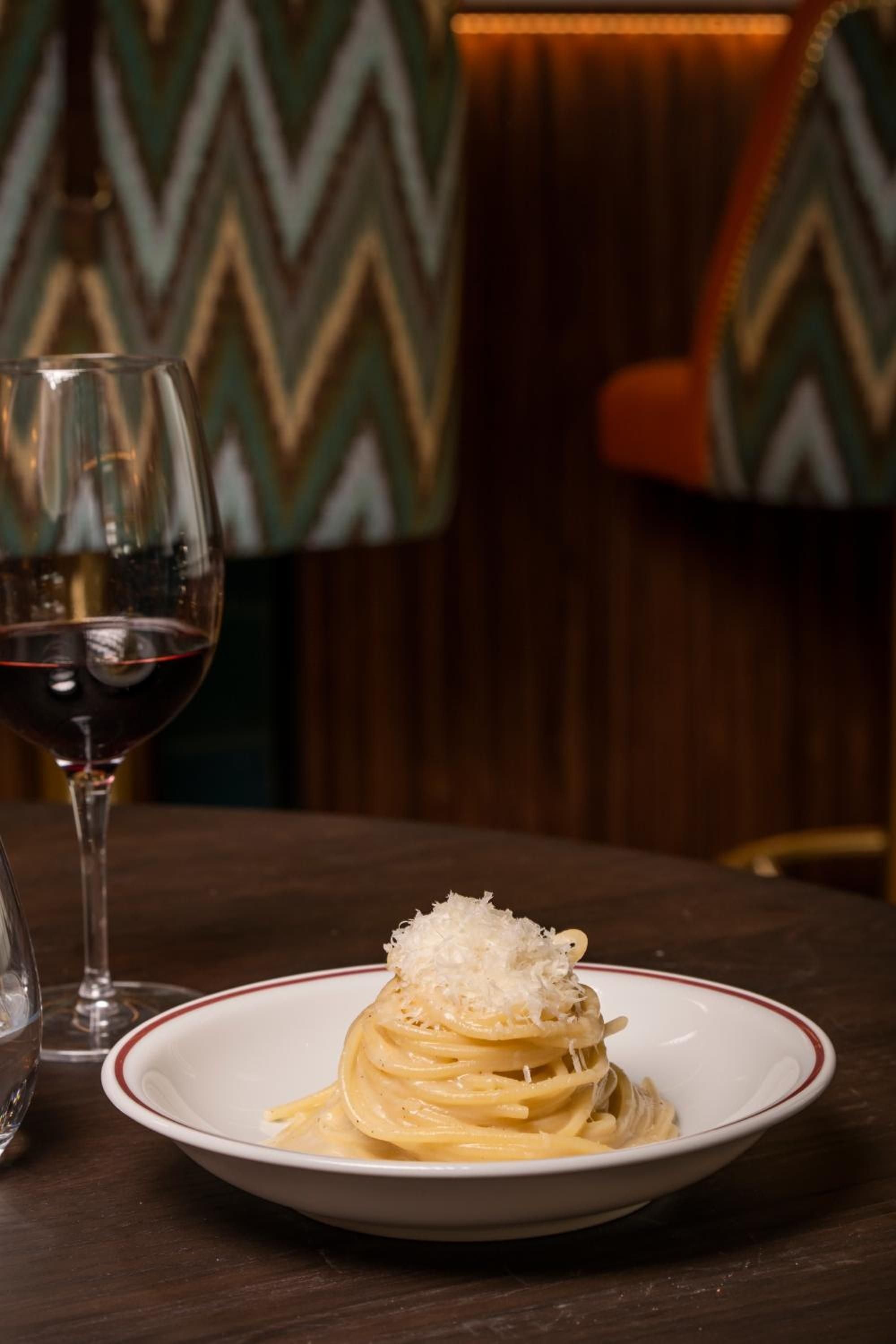 Perfectly twirled pasta crowned with delicate cheese shavings pairs with robust red wine against warm geometric wallpaper in this intimate dining moment.
