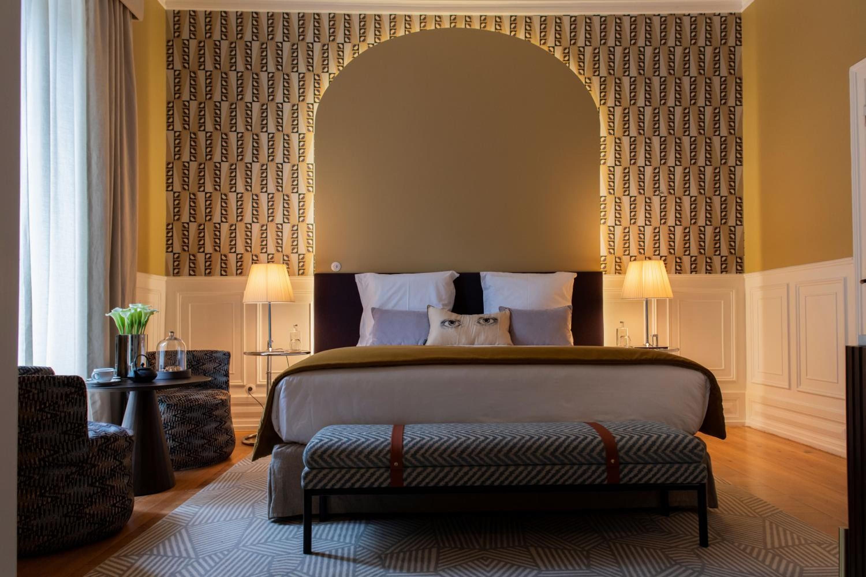 Golden light bathes the sophisticated guest room where geometric wallpaper frames an arched headboard, while thoughtful details like the whimsical cat pillow add unexpected charm to classic elegance.