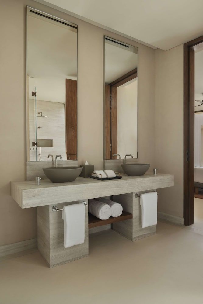 Sleek vessel sinks rest atop warm travertine vanities, where contemporary minimalism meets desert luxury in perfectly choreographed symmetry beneath frameless mirrors.