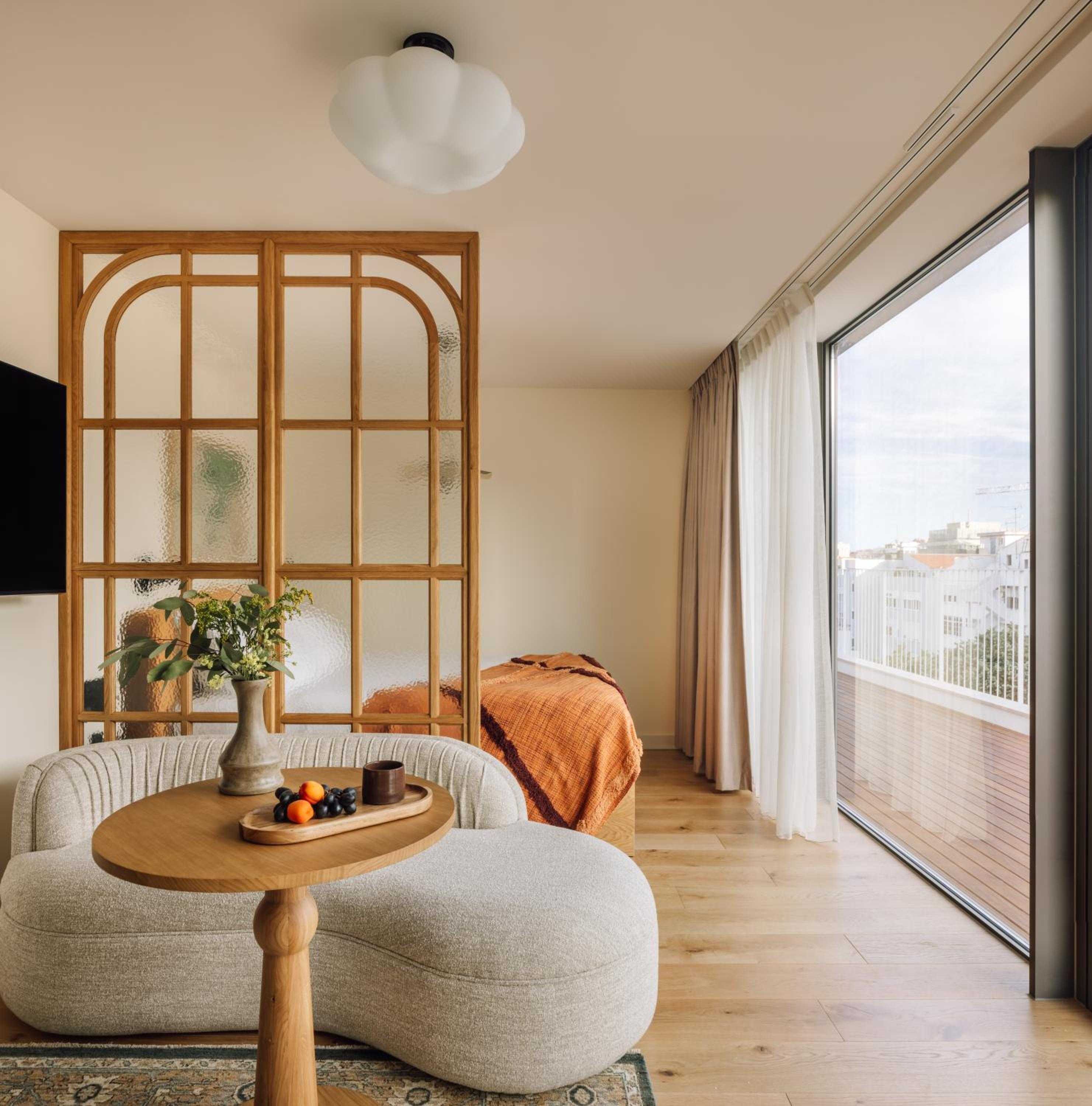 Curved seating and warm wood partitions create intimate zones in this studio apartment, where floor-to-ceiling windows frame Lisbon's terracotta rooftops beyond.