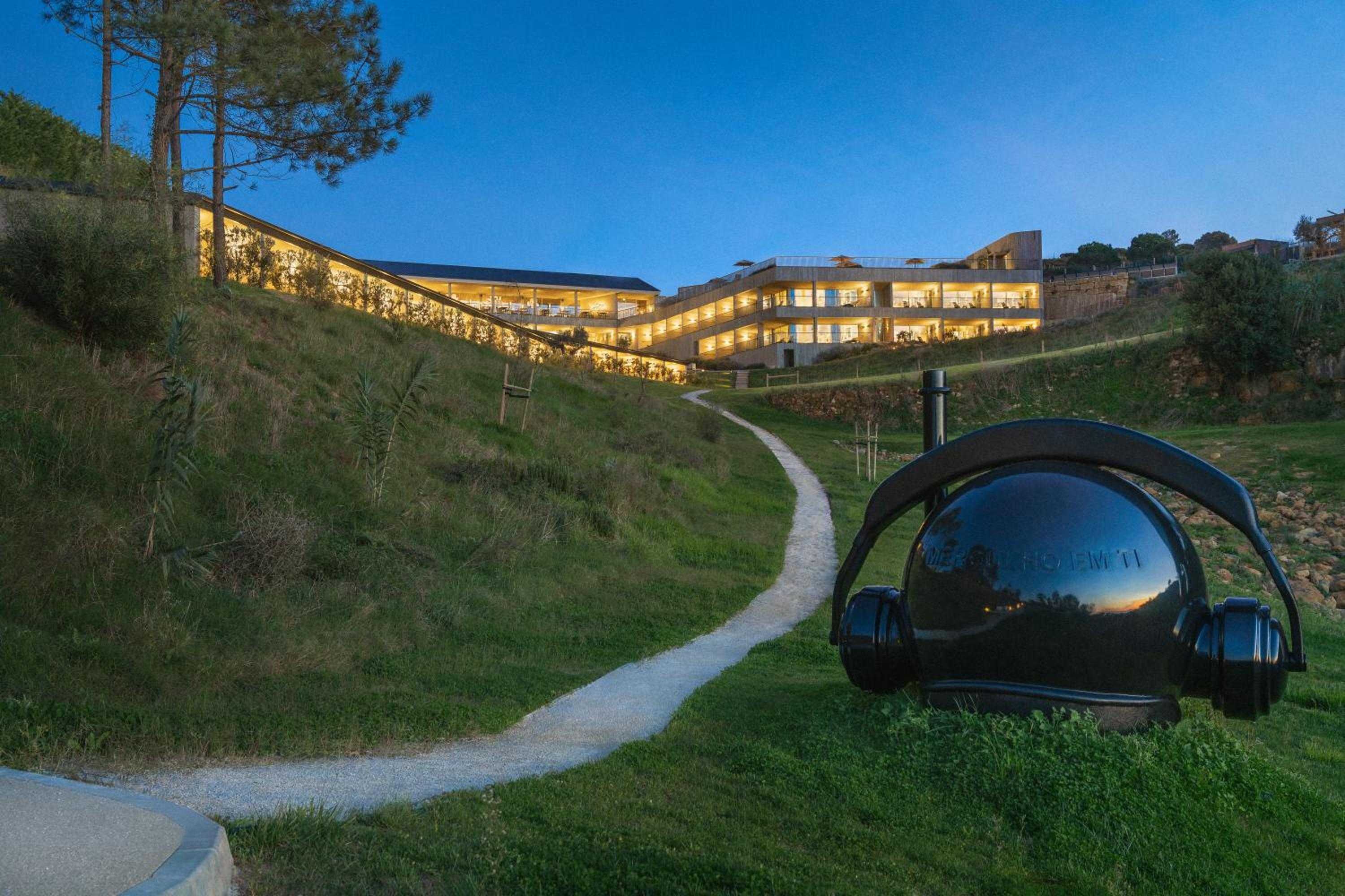 Sculptural art installations dot the hillside gardens, where winding pathways connect guests to the dramatically illuminated hotel cascading down the Mediterranean slope at dusk.