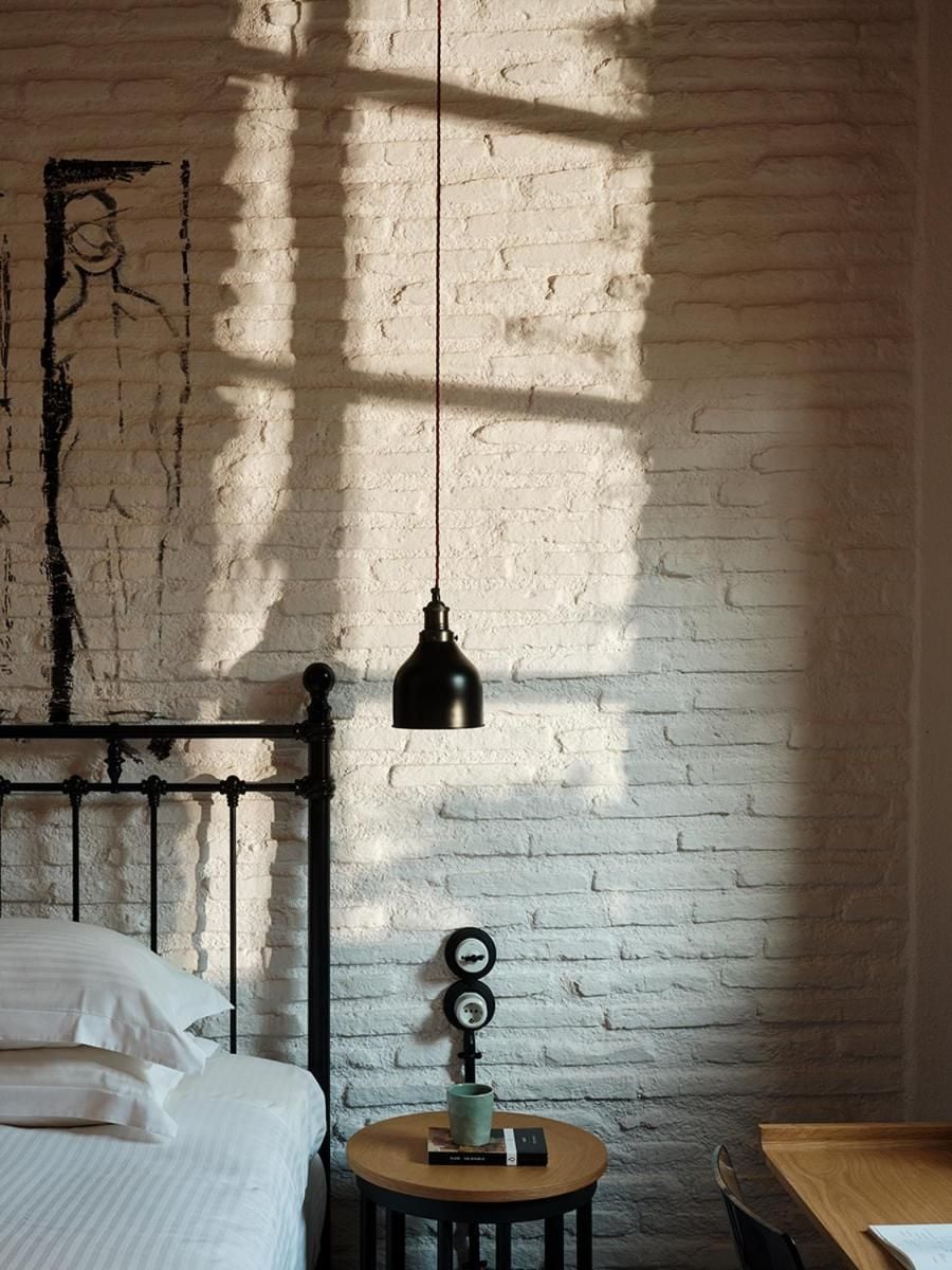 Sunlight streams through tall windows, casting geometric shadows across whitewashed brick walls where industrial pendant lighting meets vintage charm in this thoughtfully curated sleeping space.