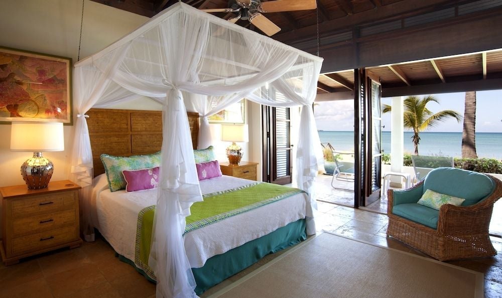 Sheer mosquito netting drapes the four-poster bed while ocean breezes flow through open doors, blending tropical comfort with barefoot luxury in this beachfront sanctuary.