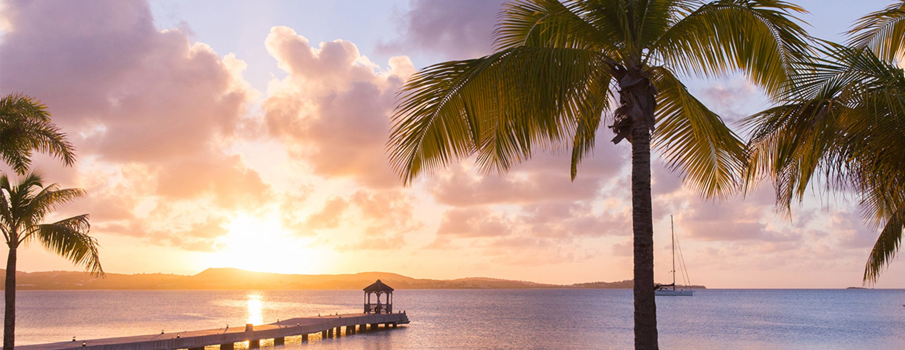 Palm fronds frame a golden Caribbean sunset as sailboats drift past Jumby Bay's weathered pier, where gentle waves lap beneath a painted sky of coral and lavender hues.