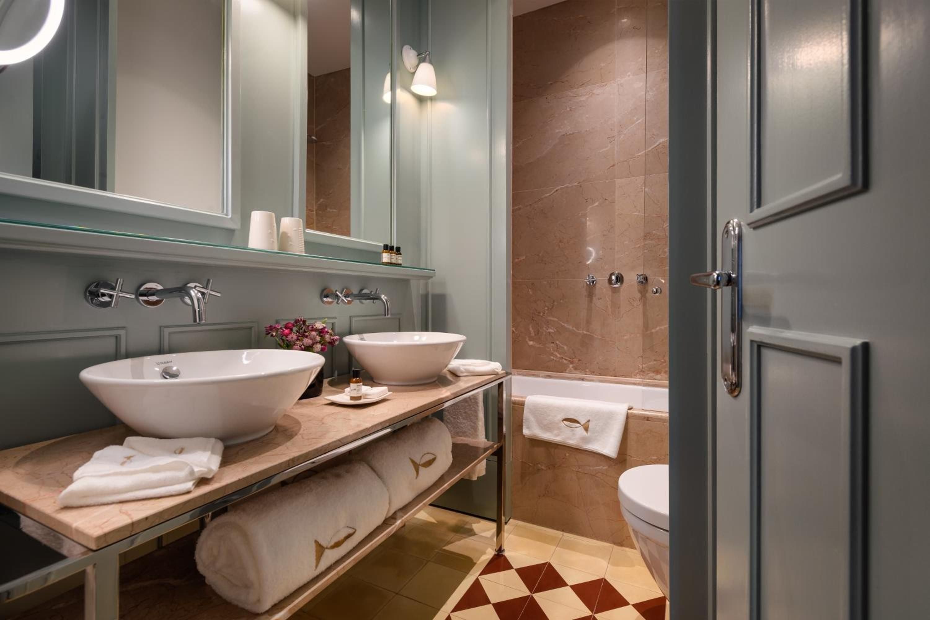 Sage-green cabinetry frames dual vessel sinks in this sophisticated bathroom, where warm marble tiling and geometric floor patterns create an intimate spa-like retreat.
