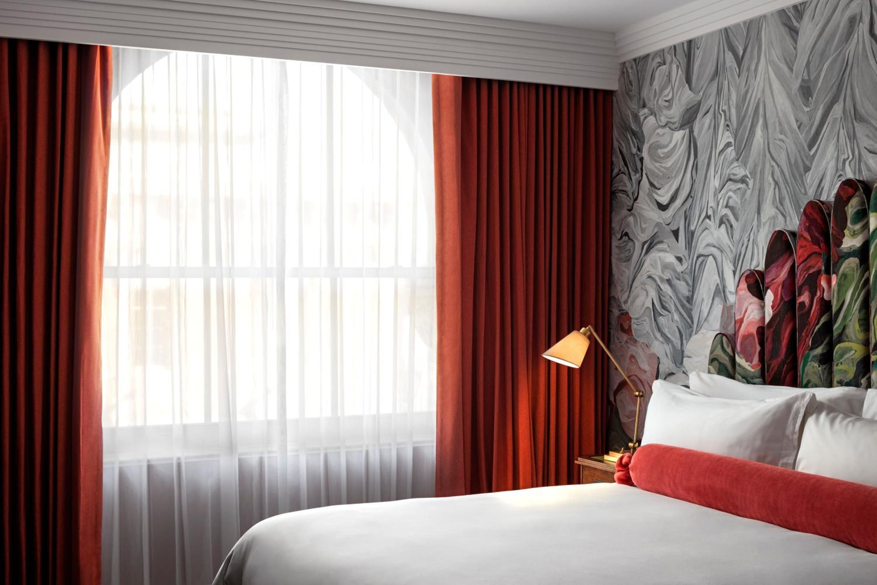 Burnt orange curtains frame city views while botanical wallpaper creates an intimate backdrop for crisp white linens and brass task lighting in this thoughtfully designed guest room.