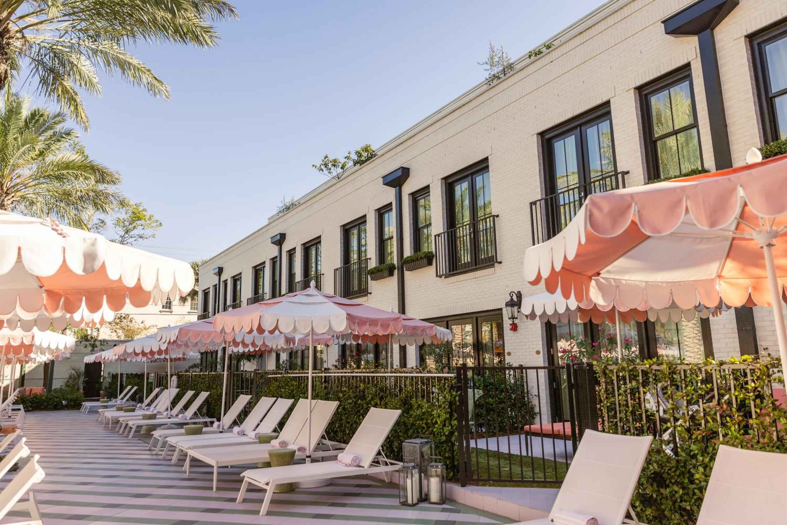 Coral-striped umbrellas cast playful shadows across pristine pool loungers, while cream brick facades and swaying palms create an effortlessly chic California retreat.