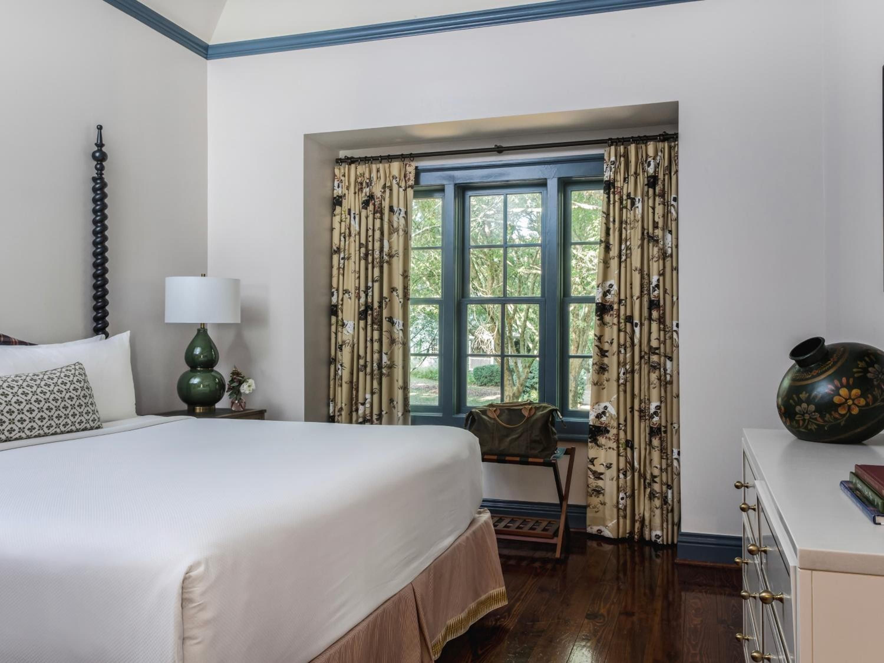 Crisp white linens contrast beautifully with rich hardwood floors and botanical curtains, while green ceramic lamps add sophisticated color to this serene guest room.