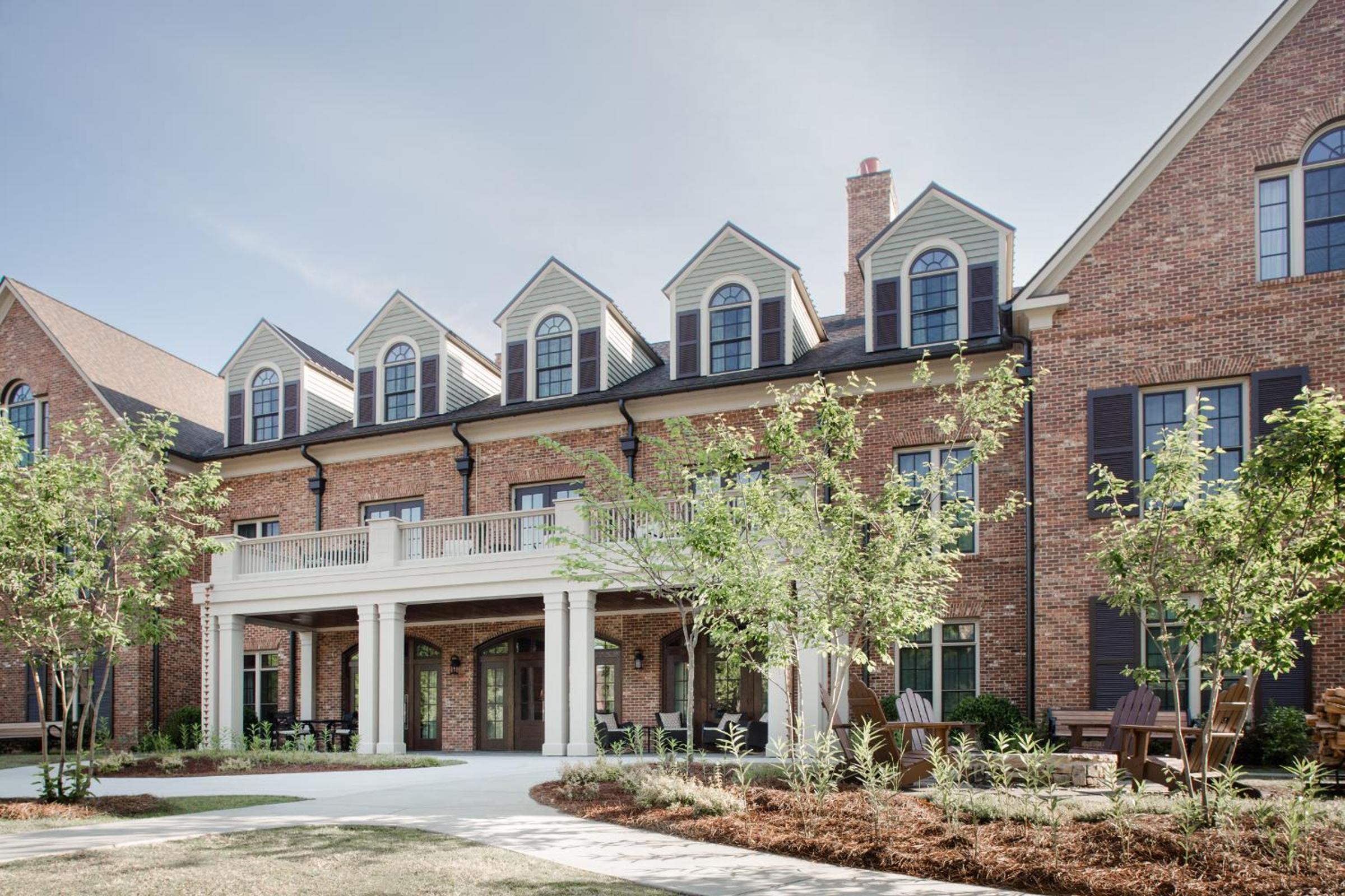 Red brick Georgian elegance meets Southern charm with white dormers punctuating the roofline, while native plantings and weathered Adirondack chairs invite lingering moments on manicured grounds.