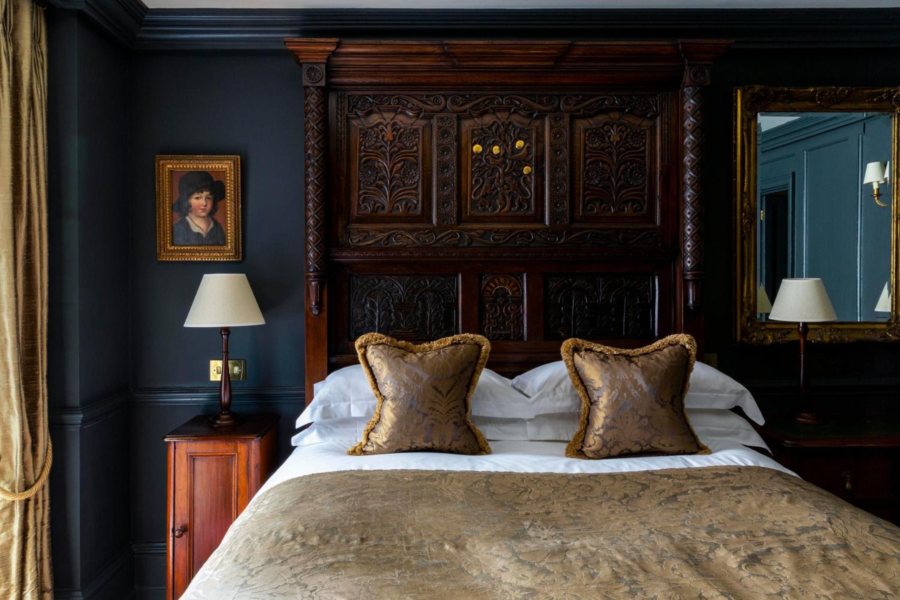 Intricately carved Jacobean paneling frames the sumptuous four-poster bed, where golden lamplight dances across navy walls and brocade pillows in this atmospheric chamber.
