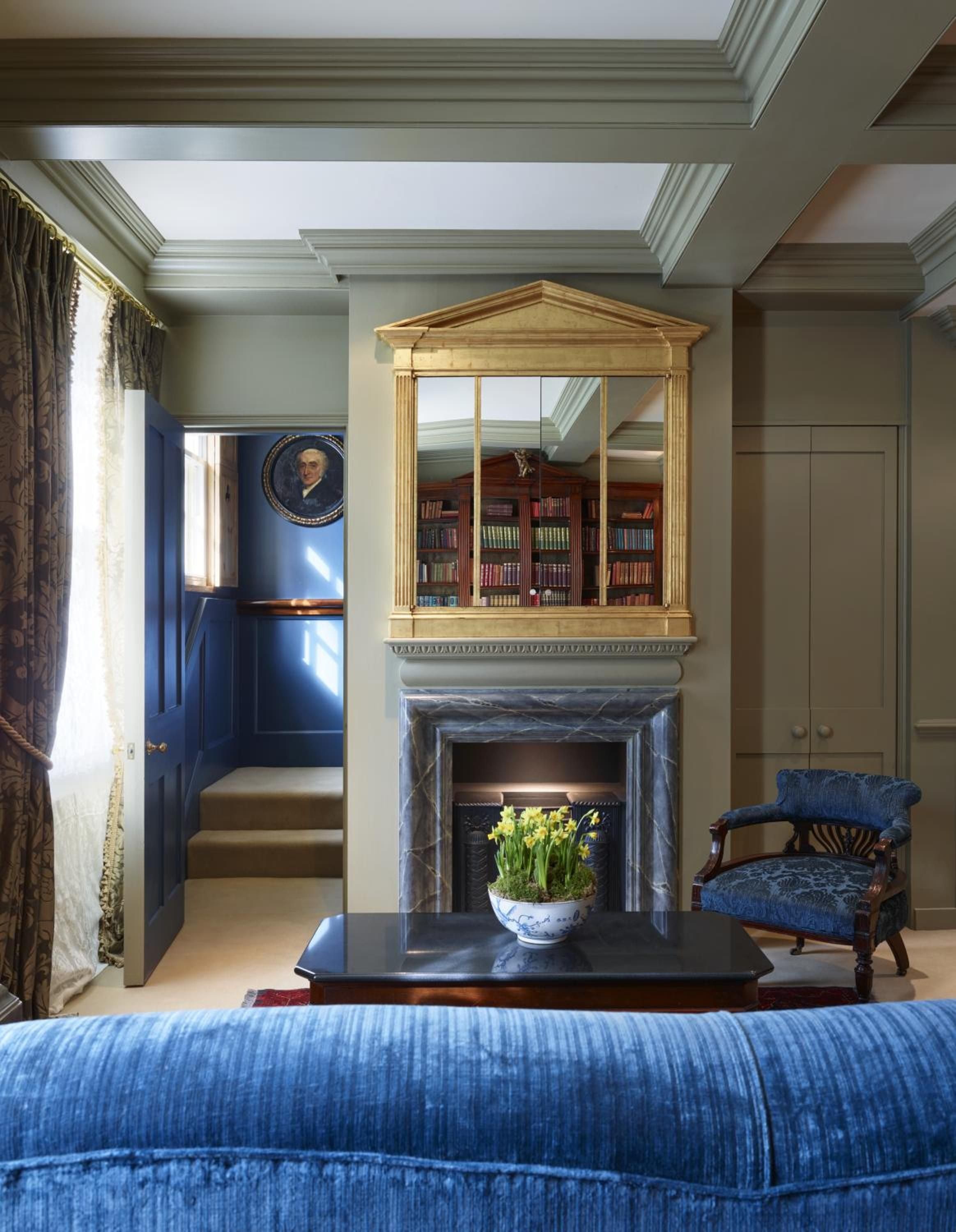 Coffered ceilings and Georgian moldings frame this intimate sitting room, where blue velvet armchairs face a marble fireplace crowned by gold-leafed library reflections.