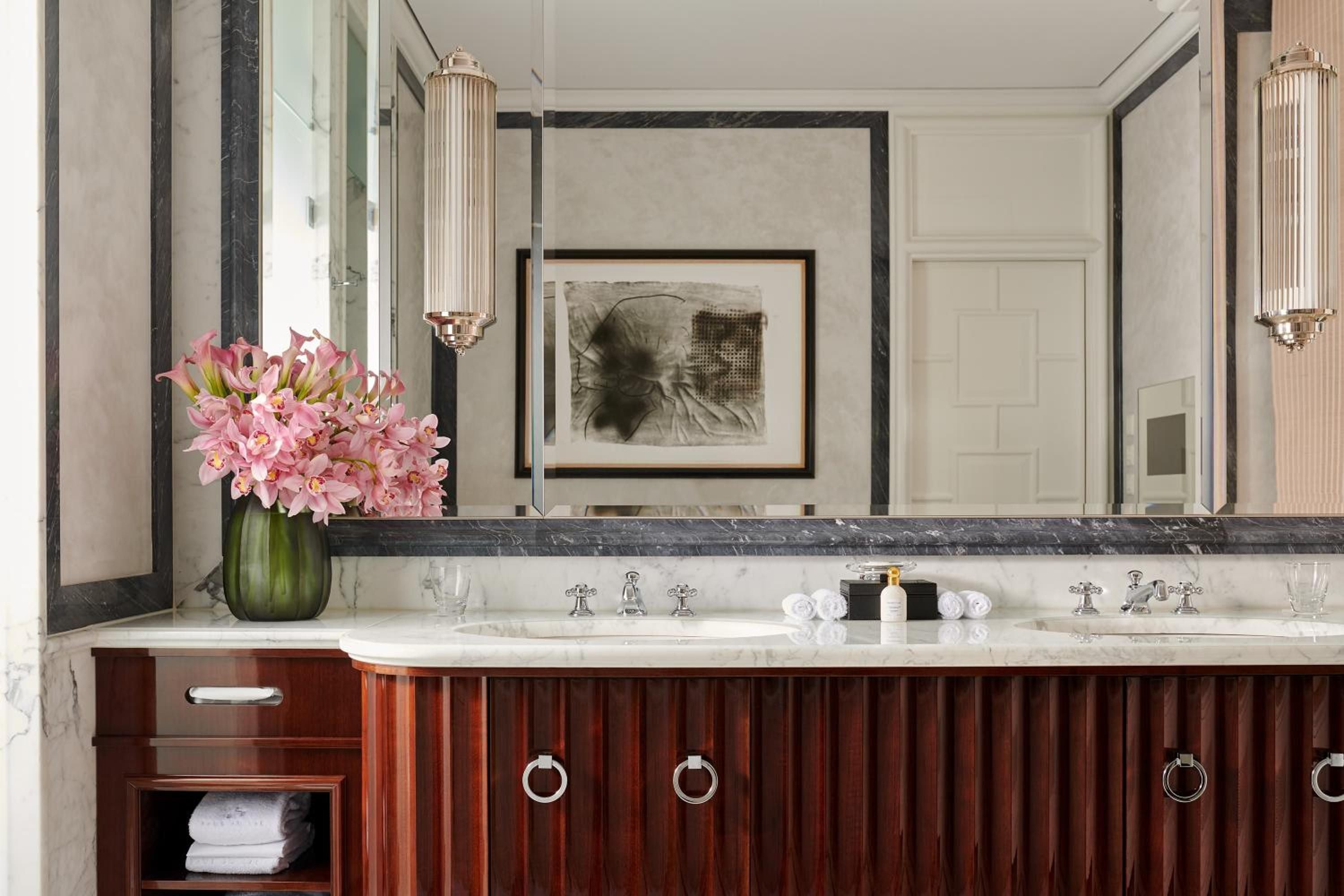 Pink orchids bloom against rich mahogany cabinetry and marble countertops, while crystal sconces cast a warm glow over polished chrome fixtures in this sophisticated vanity.