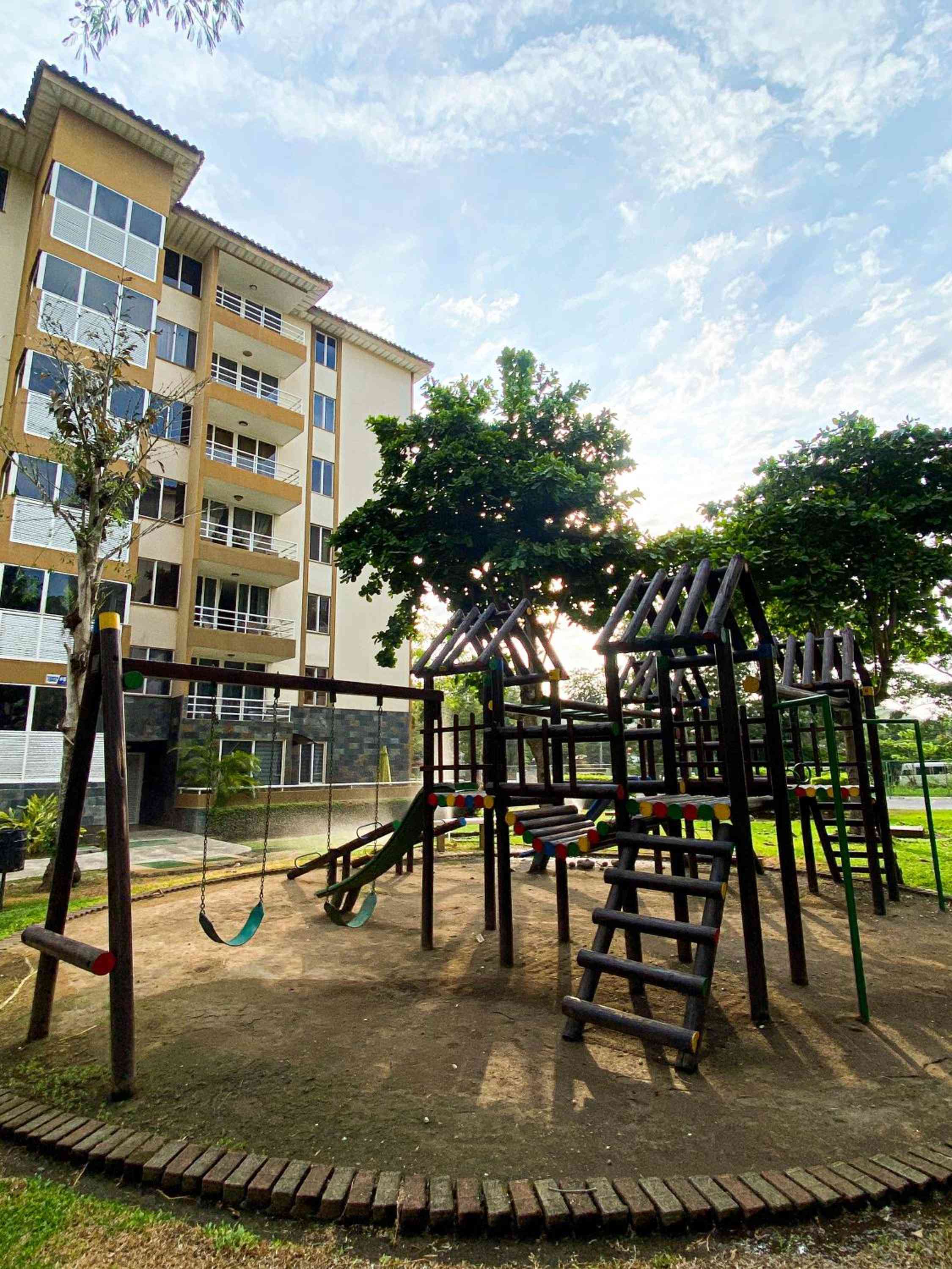 Sunlight filters through mature trees onto the children's playground, where wooden climbing structures and swings create a family-friendly sanctuary beneath the hotel's contemporary yellow facade.