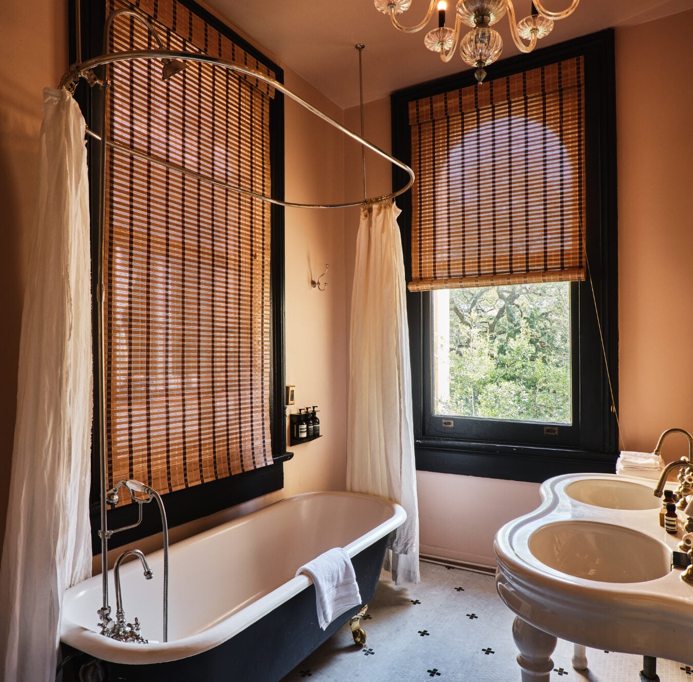 Bamboo blinds filter golden light across the clawfoot tub, where brass fixtures and white curtains create an intimate sanctuary overlooking verdant treetops.